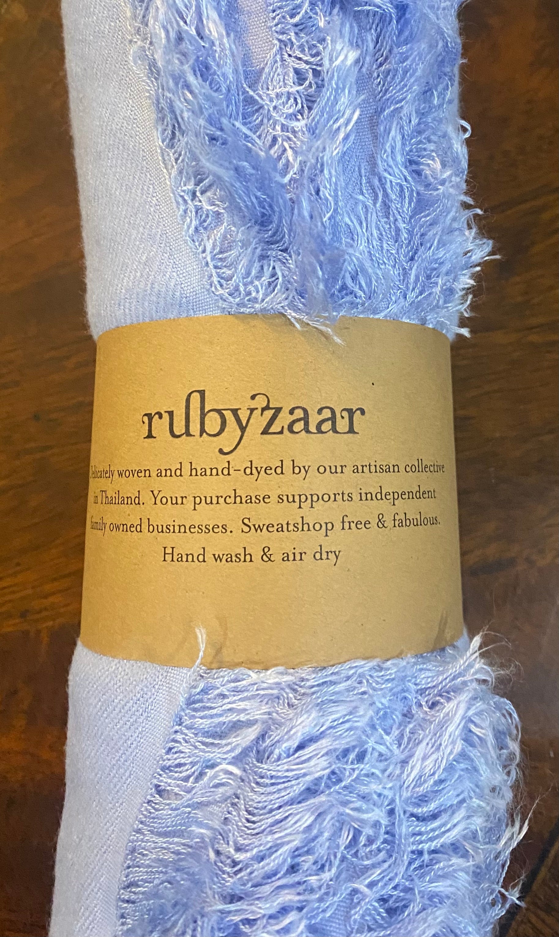 A close-up showcases a light purple woven fabric with fringed edges, wrapped in a brown paper label. The label reads "rubyzaar" and emphasizes that this fair trade, hand-woven Hima Large Scarf, Wrap, Shawl is crafted in Thailand. Promoting sustainable fashion, it’s sweatshop-free. Care instructions recommend hand washing and air drying.