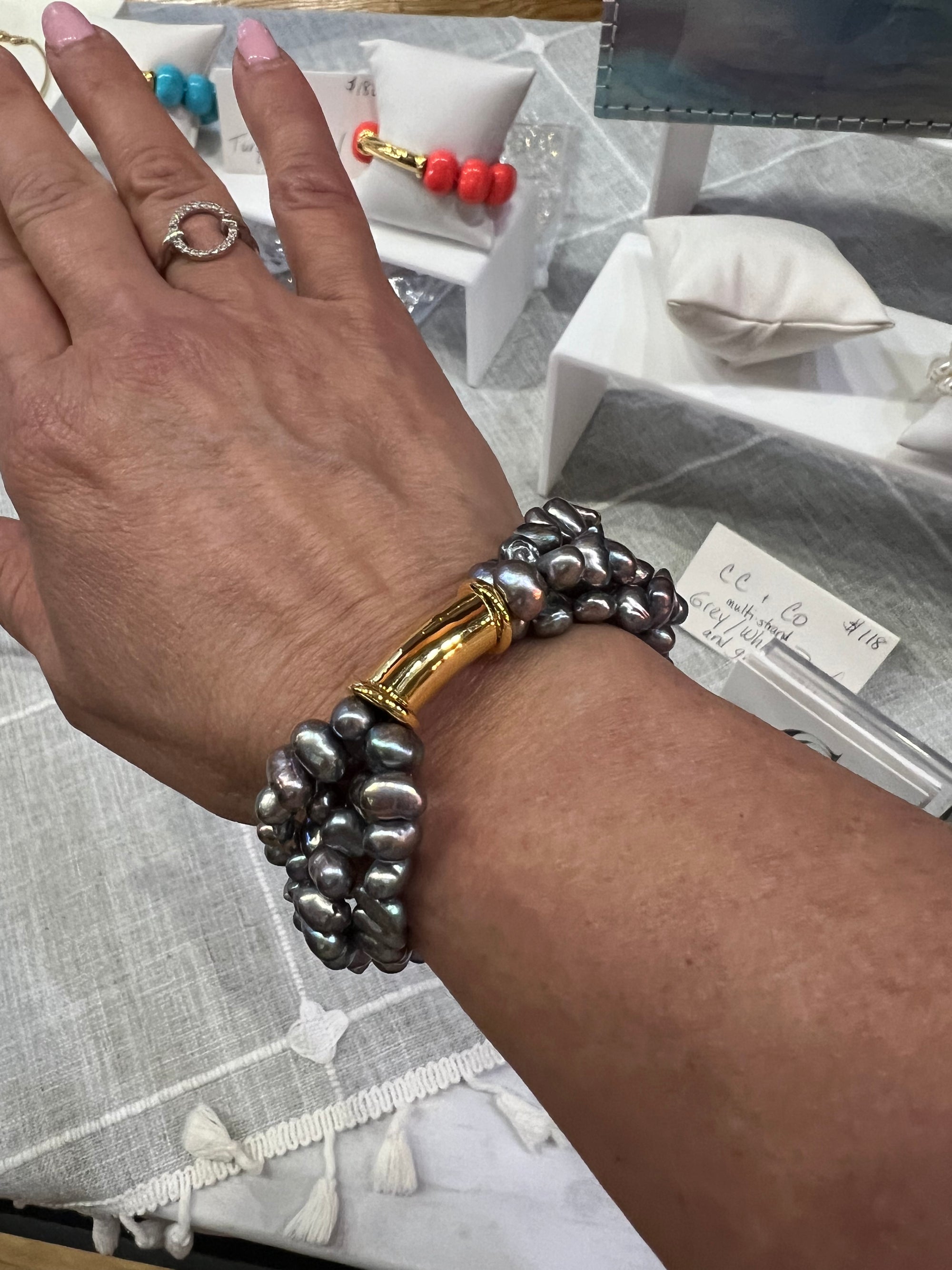 A hand wearing the Freshwater Pearl Cluster Stretchy Bracelet with Gold Hardware, featuring an iridescent gold clasp. The wrist is adorned with a ring, while vibrant beaded bracelets stretch across cushioned setups on a light-colored tablecloth in the background.