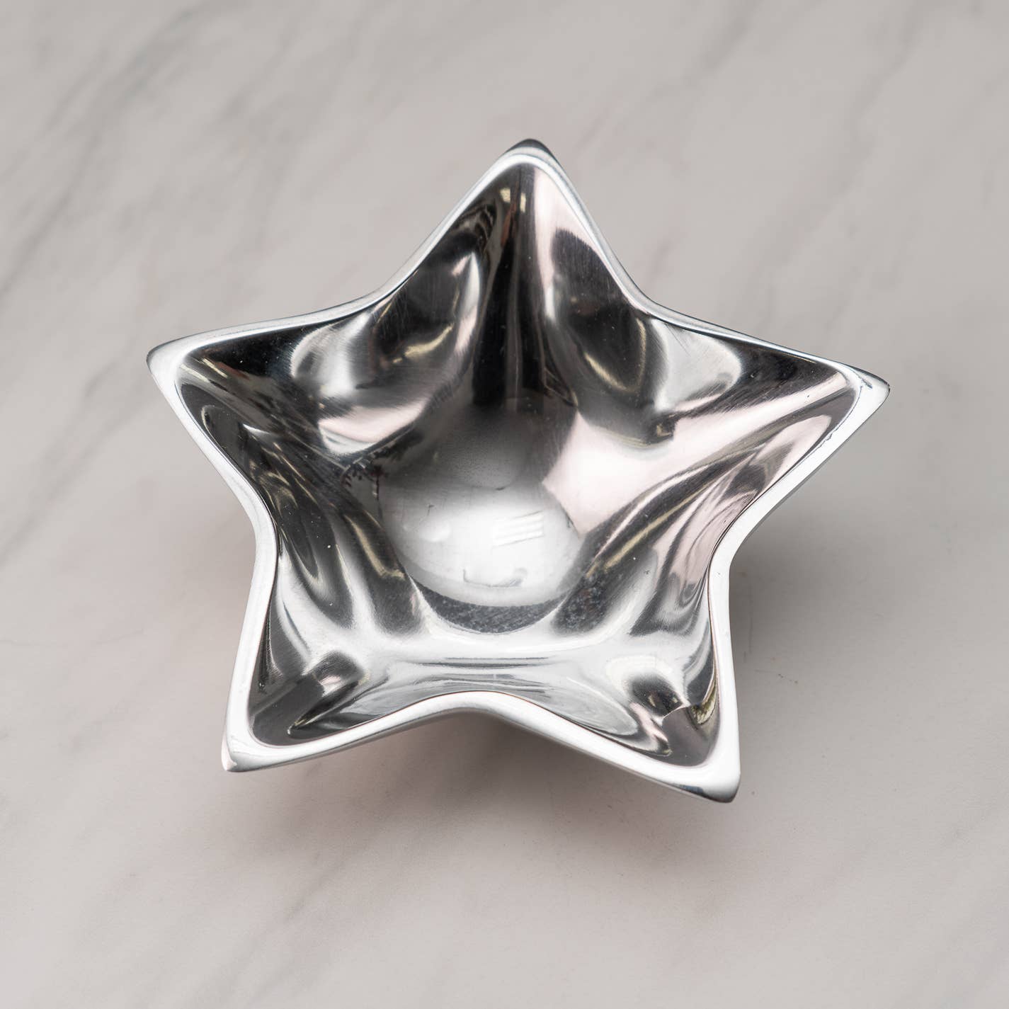 An Aluminum Star Tray with a sleek, silver star-shaped design rests on a light gray marble surface. Made from food-safe aluminum, its reflective finish captures the light beautifully, emphasizing its smooth curves and glossy appearance, which lend it a modern and decorative look.
