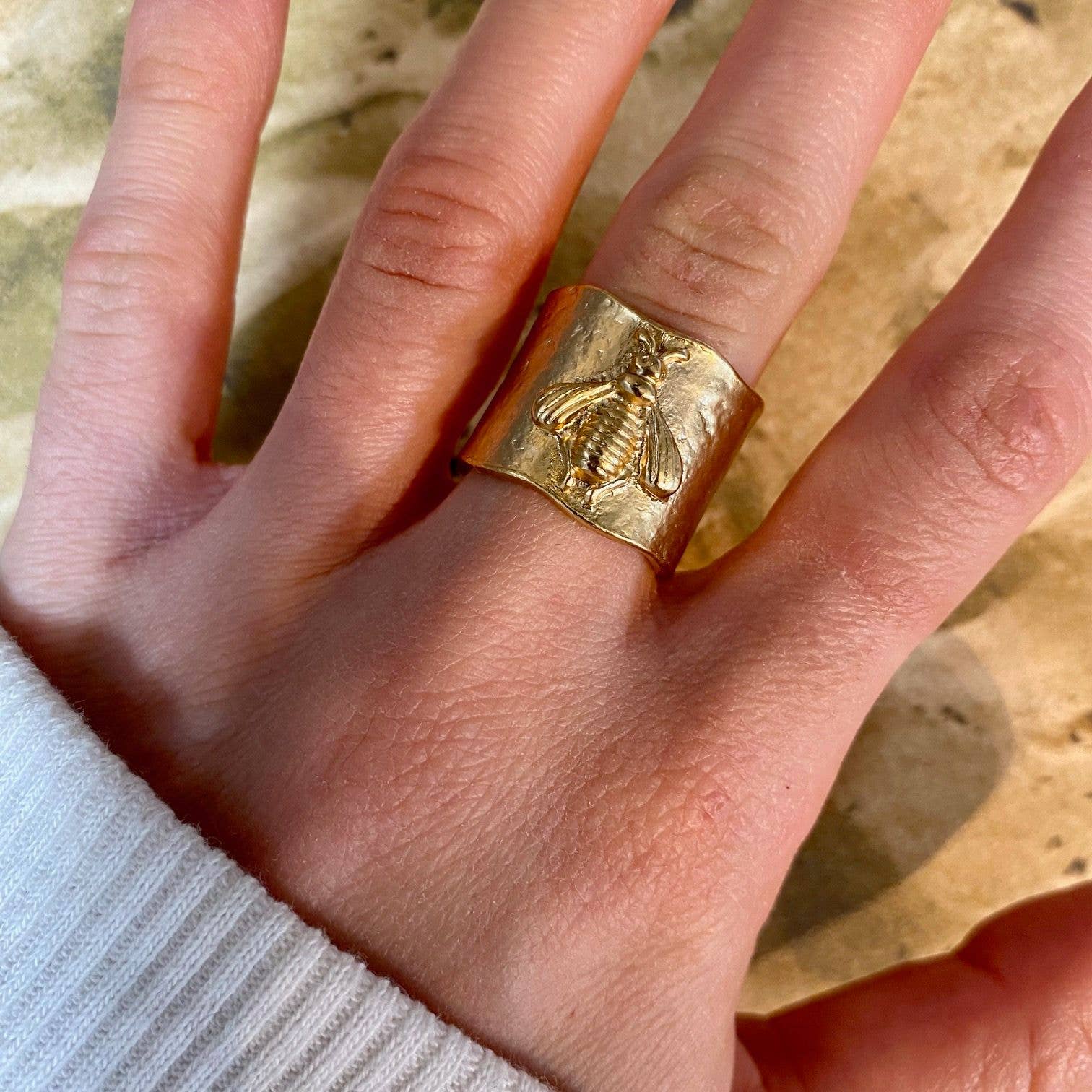 A hand with a white sleeve visible is wearing the Bumblebee Ring: Gold on the middle finger, featuring a detailed bee design. It's crafted in 24 kt gold and part of our exclusive Bee Collection, set against a textured, earth-toned background.