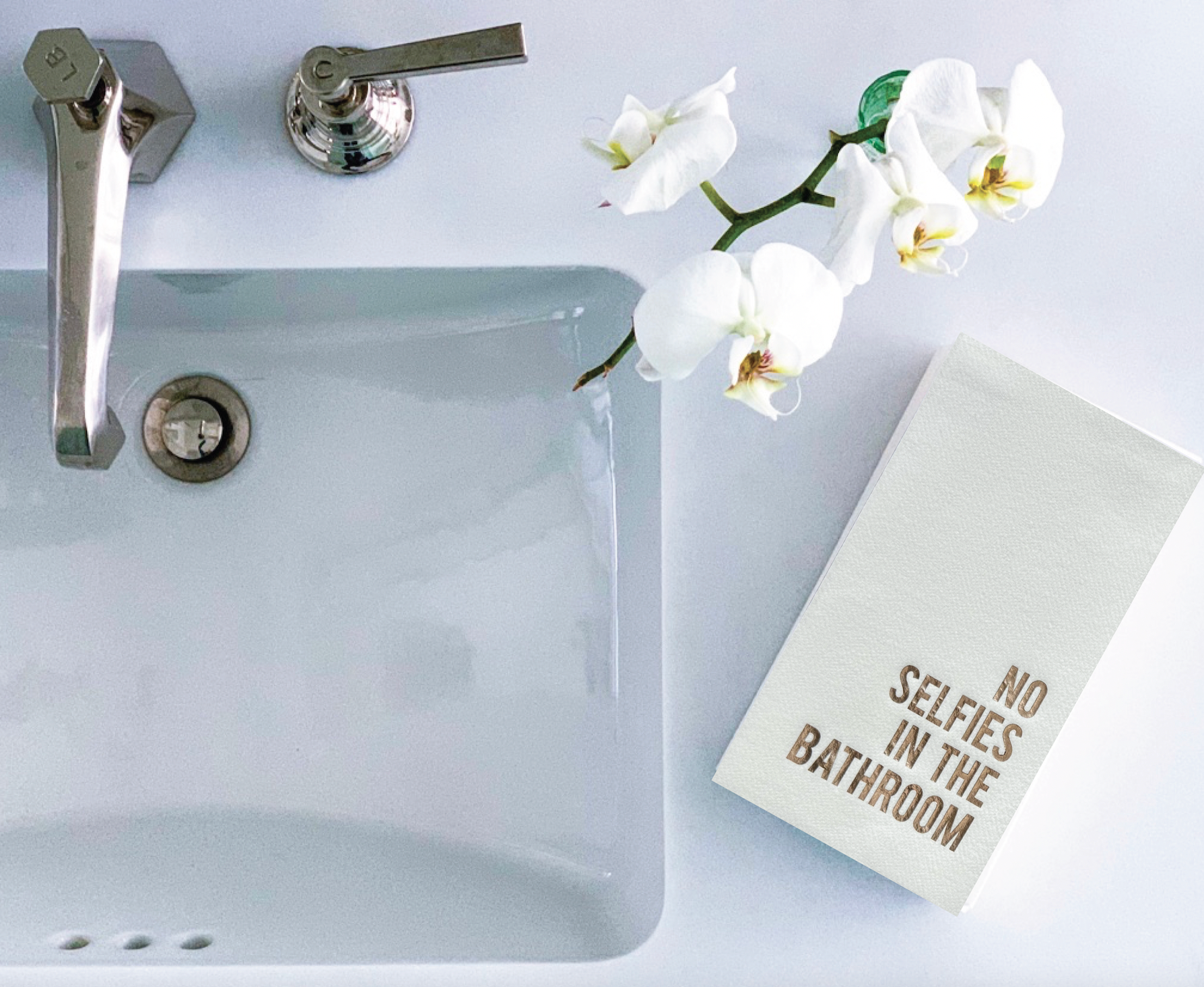A bathroom sink with a contemporary silver faucet is complemented by a white orchid on the counter. Positioned to the right, a stack of Cloth-Like Guest Towels - Year Round Themes displays elegant gold lettering that says, "NO SELFIES IN THE BATHROOM." The overall ambiance is clean and minimalist.