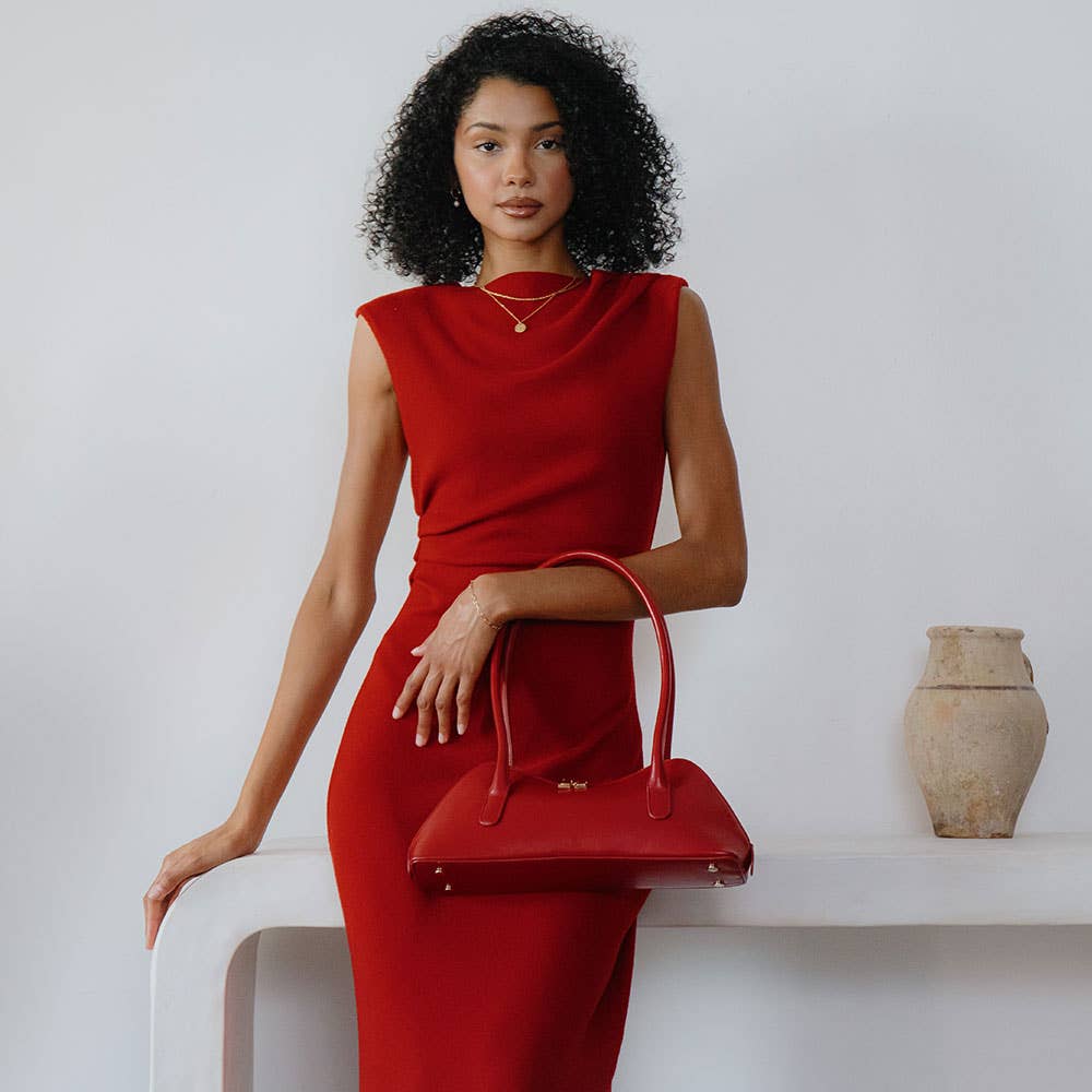 A curly-haired woman stands against a white background wearing a sleeveless red dress, holding a Dakota Recycled Vegan Shoulder Bag. Behind her, a simple white shelf holds a beige ceramic vase. The minimal and elegant setting is enhanced by the bag's OEKO-TEX certified lining.