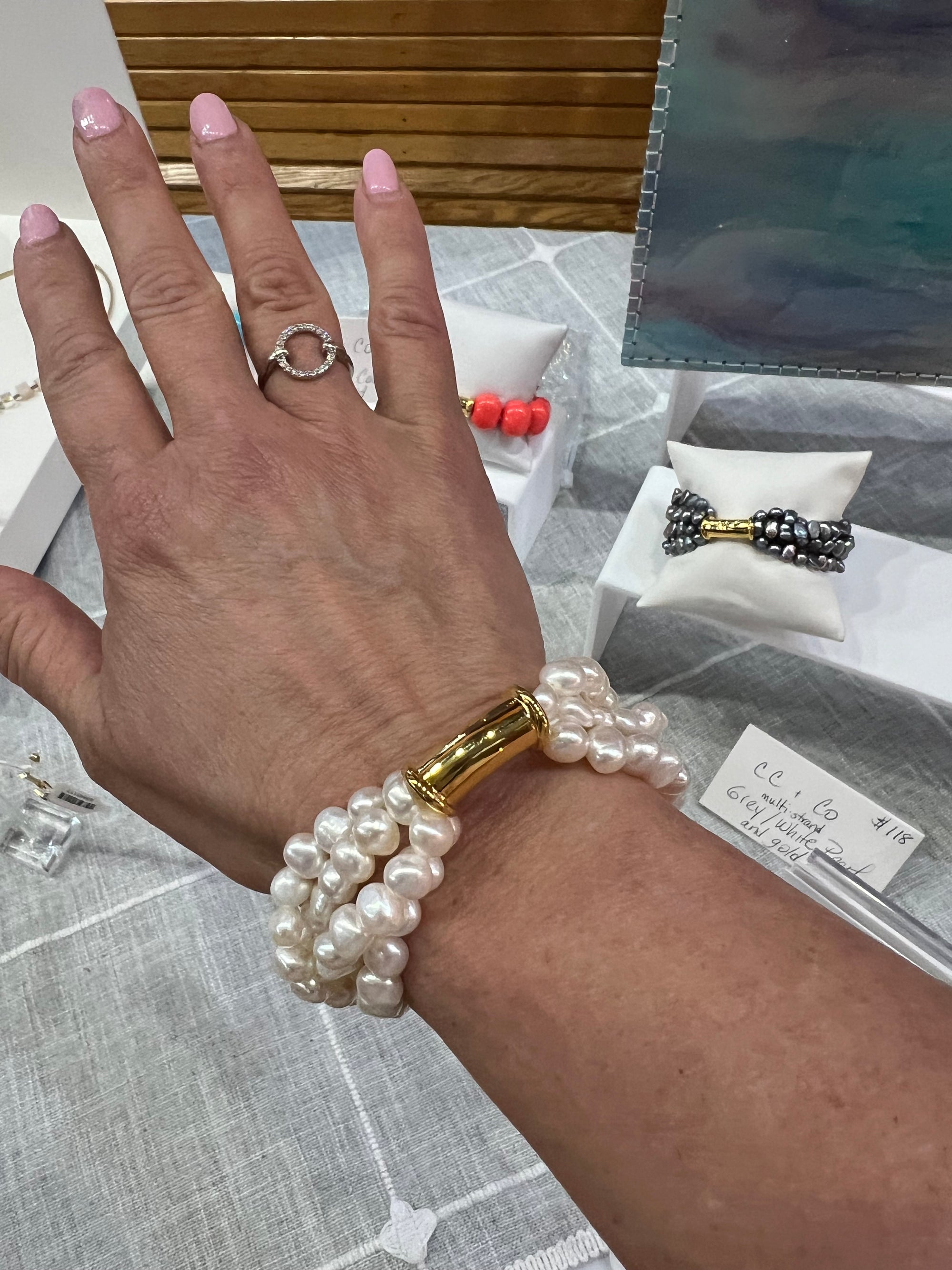 A hand with pink nails displays the Freshwater Pearl Cluster Stretchy Bracelet with its iridescent pearls and gold hardware. The hand also wears a silver ring. In the background, jewelry displays featuring bracelets and rings, some adorned with pearls and colorful beads, are set on a light gray cloth.