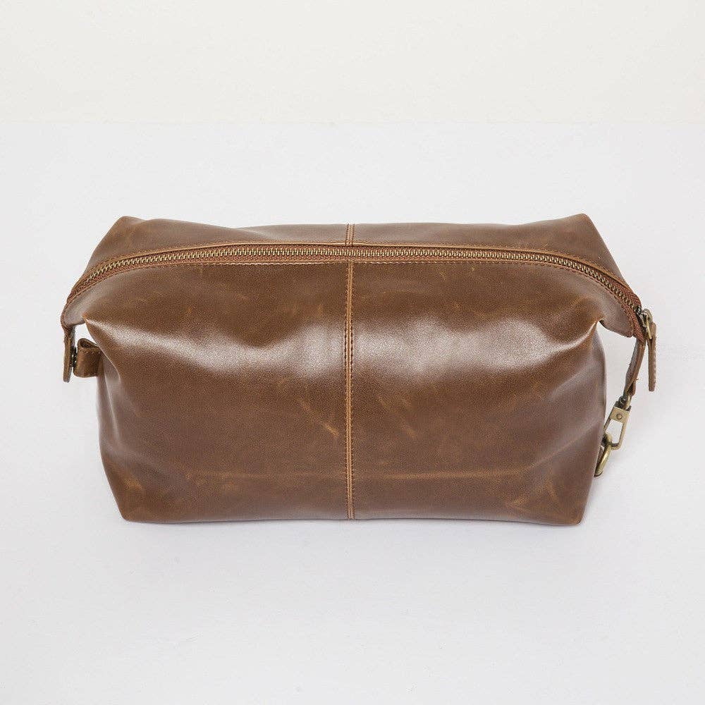 The Journeyman Dopp Kit is made from brown vegan leather, featuring a zipper along the top. It has a straightforward, rectangular design with visible stitching and rounded edges. The smooth, slightly worn texture of the leather adds vintage appeal, complemented by a metal zipper pull—ideal for your grooming essentials.