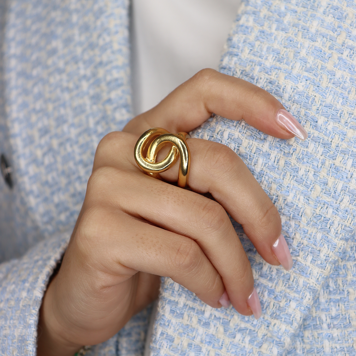 A hand with manicured nails holds the lapels of a light blue textured jacket, wearing an Intertwined Link Oversize Ring made of 24-kt gold, showcasing an abstract interlocking design on the index finger. The background is blurred to focus on the hand and ring.