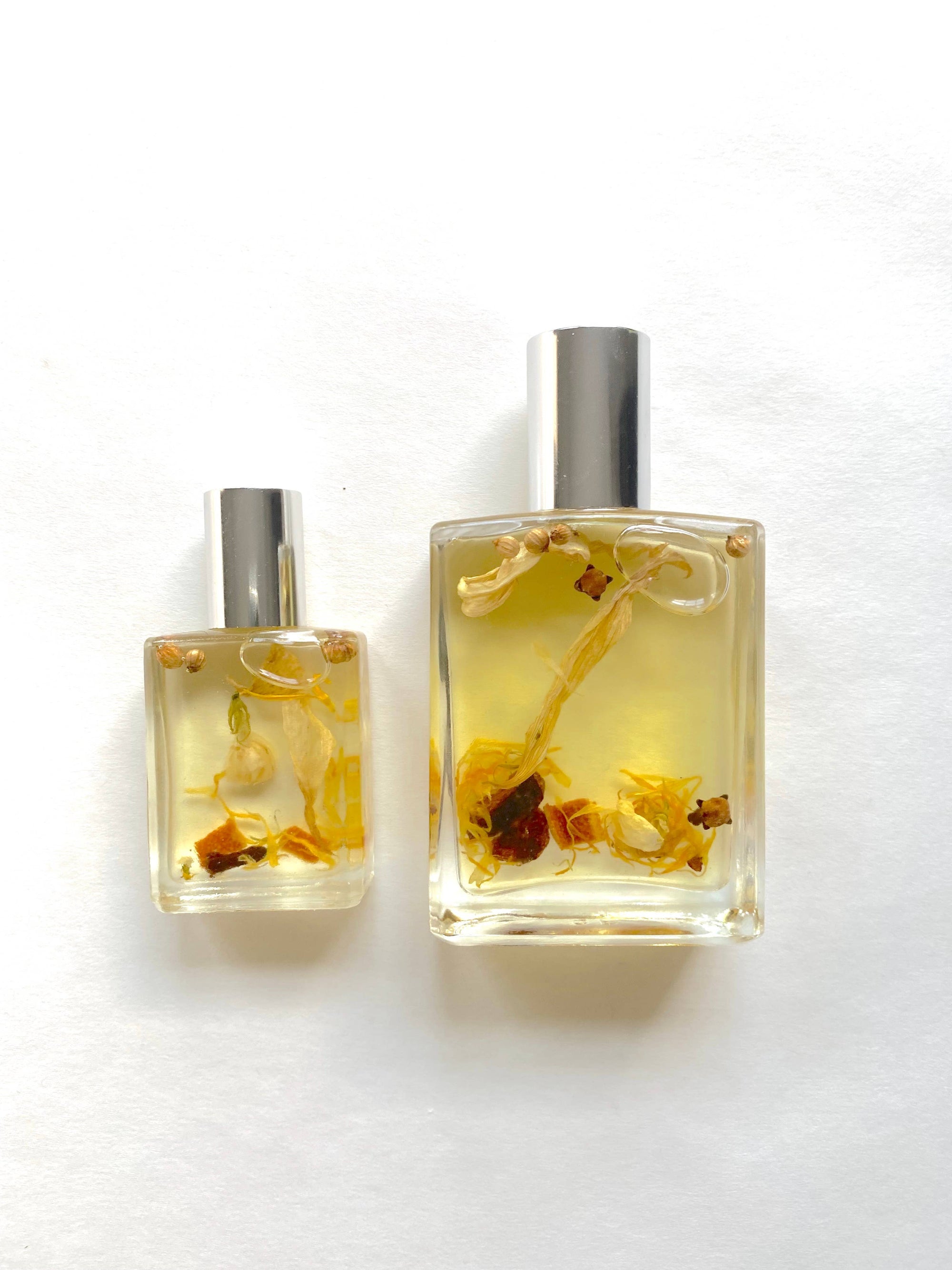 Two bottles of "Need Sleep" Aromatherapy Body Oil, crafted from glass with elegant silver caps, are filled with a soothing light yellow liquid enriched with a therapeutic fragrance. The larger bottle is positioned on the right, and both display dried botanical elements like petals and stems suspended within. They are set against a pristine white background.