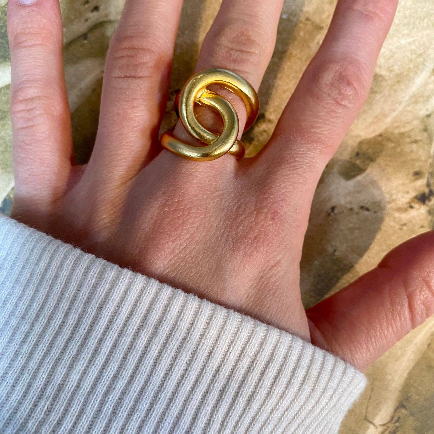 A hand with a beige sleeve elegantly displays the Intertwined link oversize ring on the middle finger. Crafted from 24-kt gold, this ring boasts a modern artistic design featuring two overlapping loops. It is set against a textured, marbled surface in shades of brown and tan.