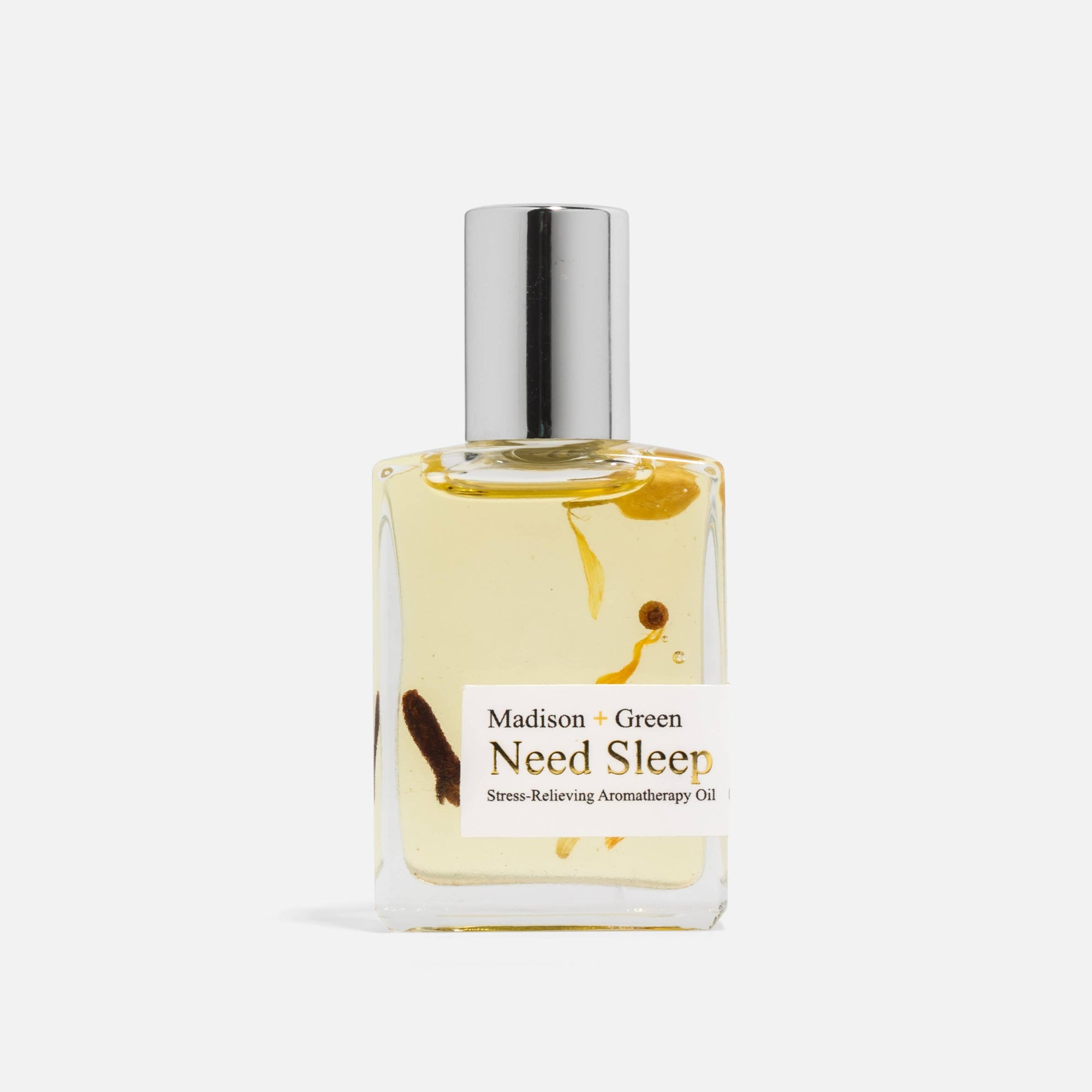 A glass bottle of the "Need Sleep" Aromatherapy Body Oil from Madison + Green is set against a plain white background. This bottle, filled with light yellow organic essential oils and visible herbal fragments, is topped with a silver cap and features a white label with black text that promises therapeutic aromatic relief.