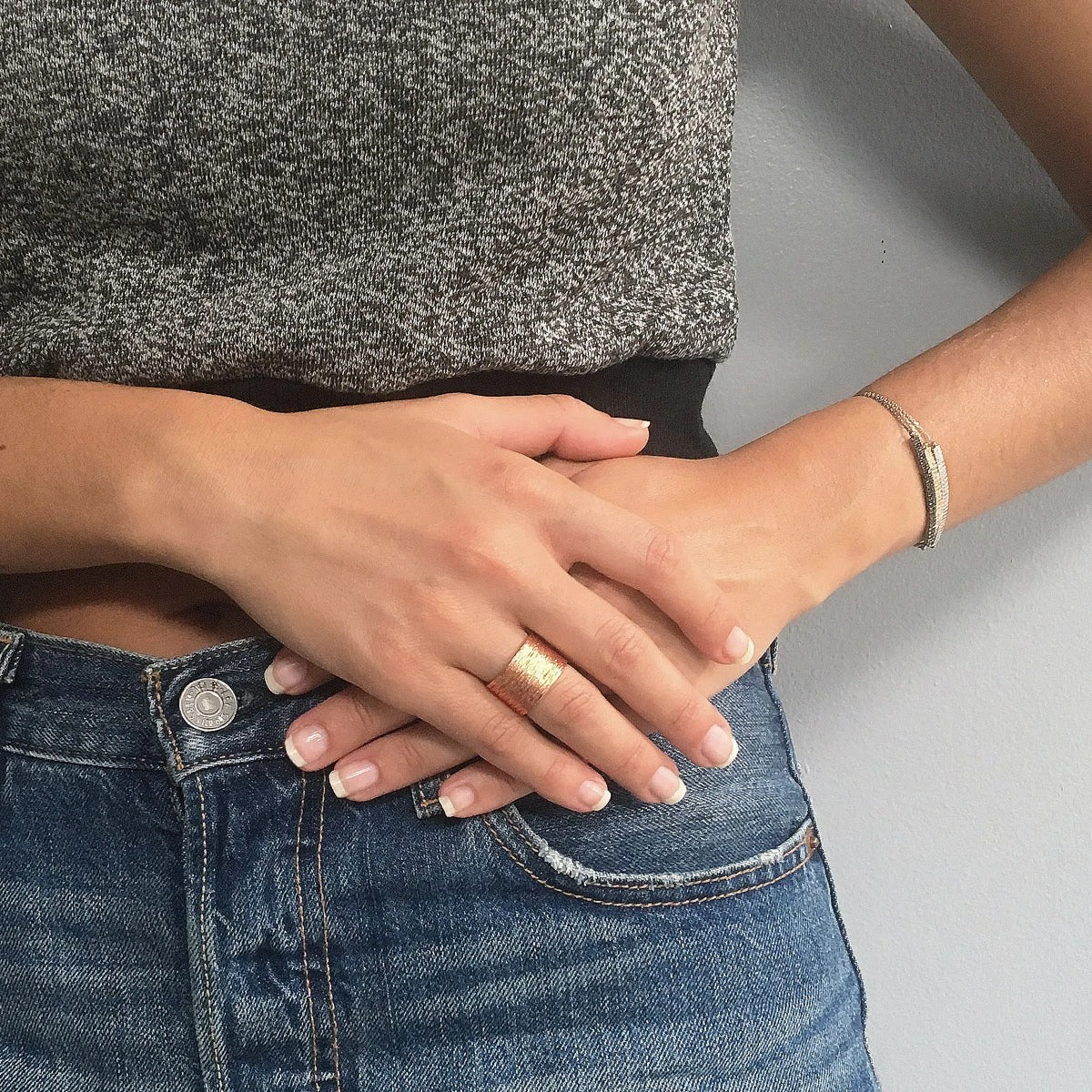 A person wearing a gray top and blue jeans stands against a light gray background. Their hands are crossed at the waist, showcasing neatly manicured nails adorned with a minimalist Brushed Ring and a thin bracelet. The copper ring is enhanced with 24 kt gold plating. The jeans feature a silver button and visible pocket stitching.