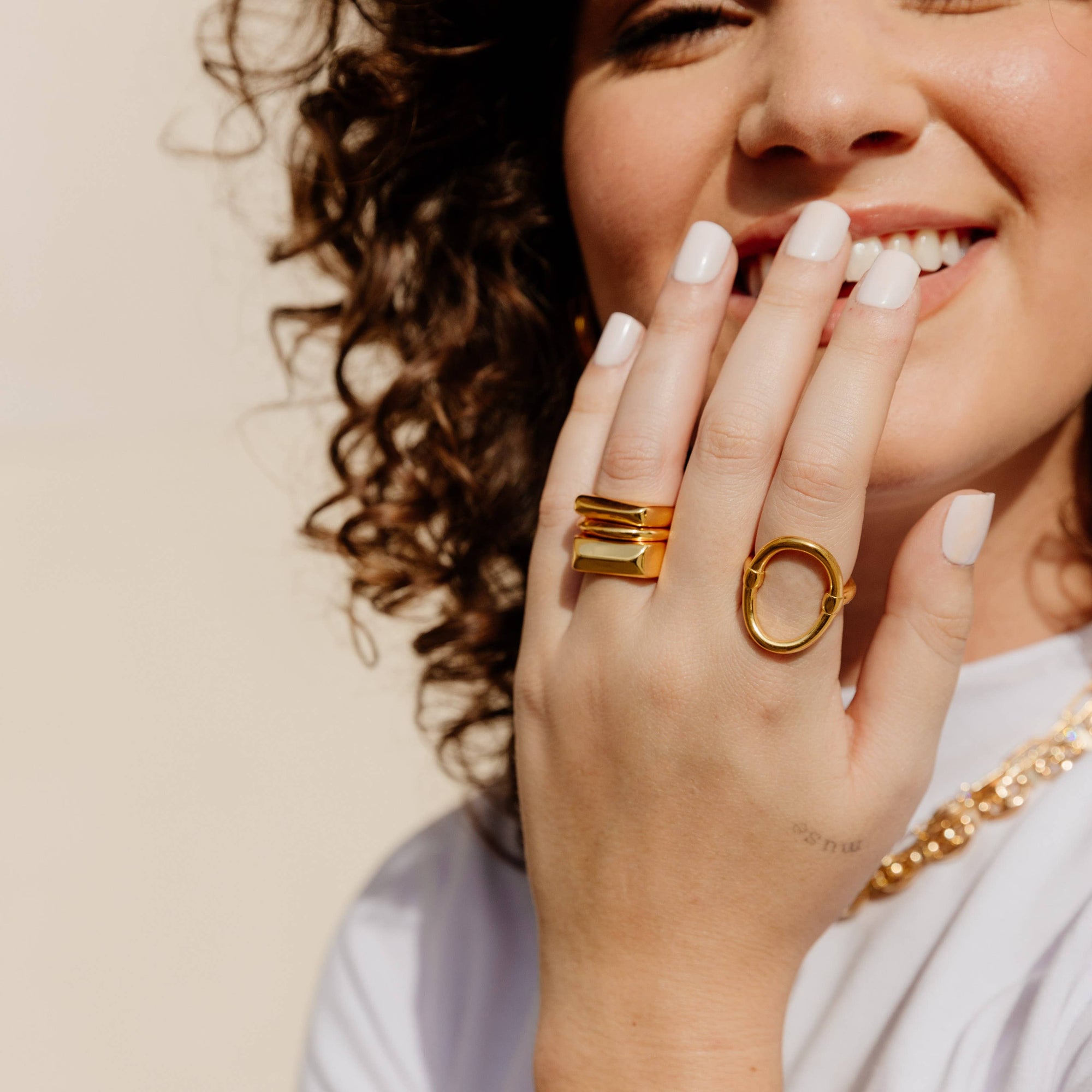 A person with curly hair smiles as they touch their face, wearing a white shirt and accessorizing with elegant jewelry in 18k gold plating. Among the pieces is the stunning Nova Ring Set, showcasing beautifully stacked rings, including one standout ring with a large oval design. Their nails are painted white, contrasting against the plain, neutral background.