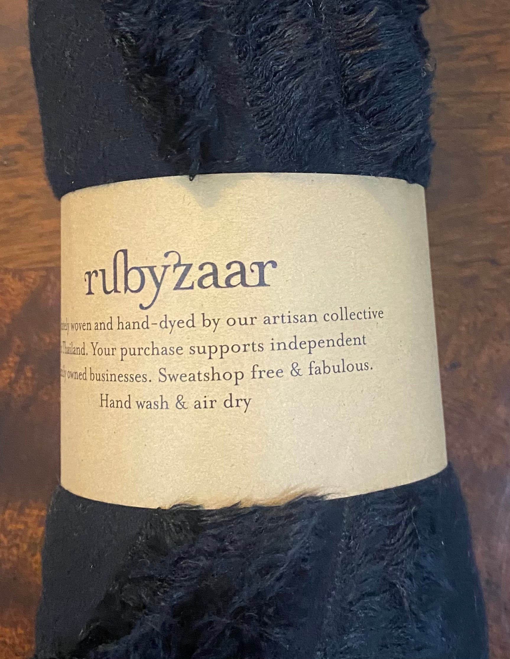 A soft, dark fabric roll wrapped with a brown label that reads: "rubyzzaar, Hand woven and hand-dyed by our artisan collective in Thailand. Your fair trade purchase supports independent women-owned businesses. Sweatshop free & fabulous. Hand wash & air dry." This is the Hima Large Scarf, Wrap, Shawl.