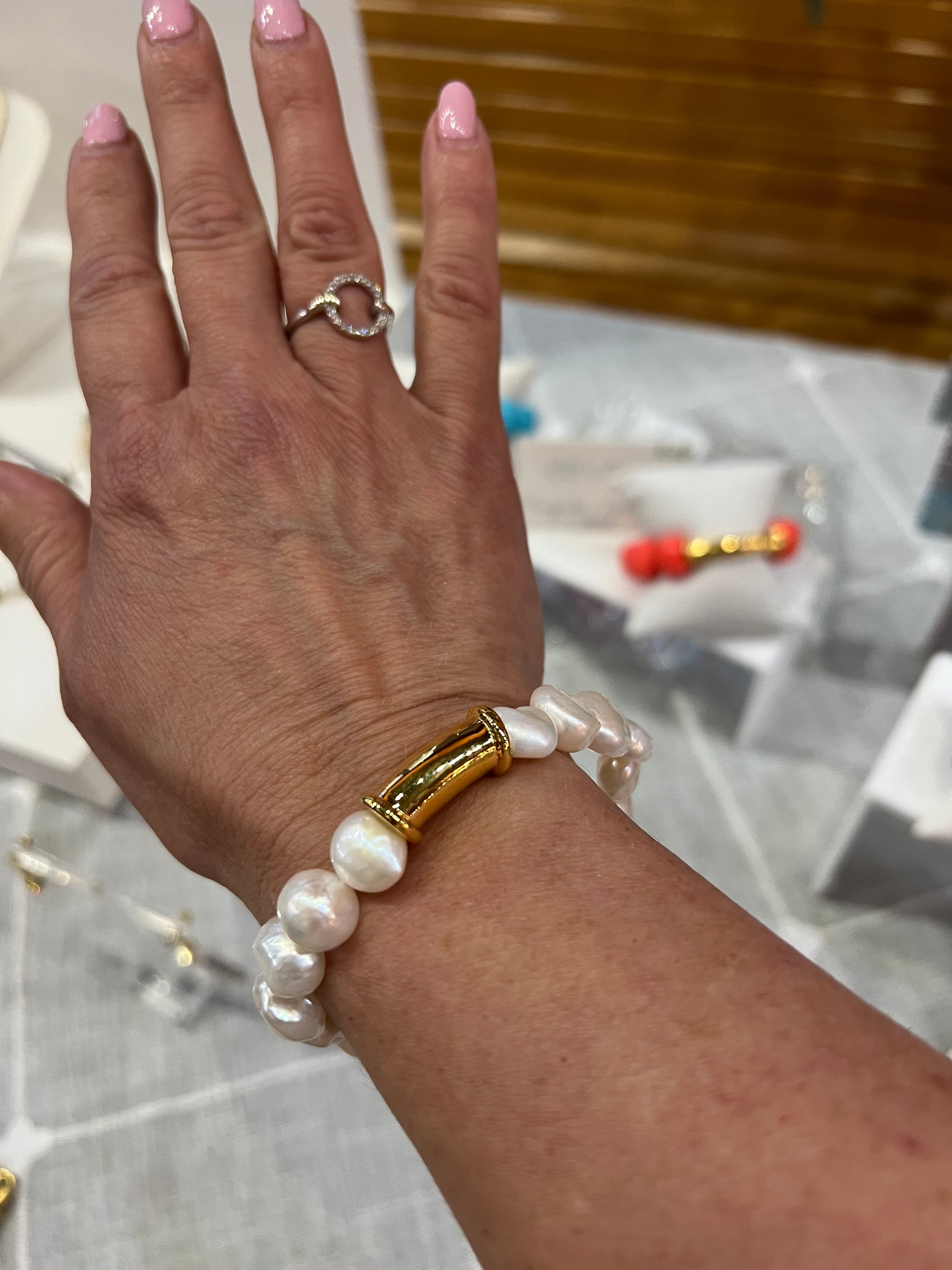 A hand with light skin is adorned with the Nugget Freshwater Pearl Bracelet, featuring a central 14-karat gold cylindrical accent. The person also wears a silver ring with a circular design on their ring finger. Their nails are polished in pale pink nail polish, set against a blurred jewelry display backdrop.