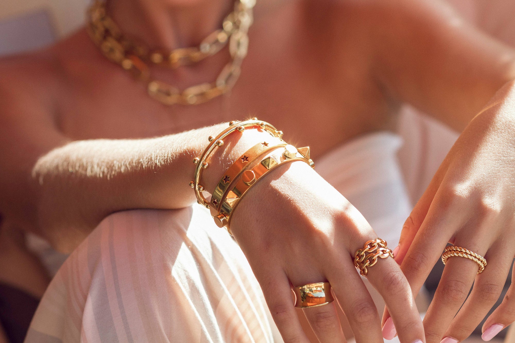 The woman is adorned with 18k gold plated bracelets and rings. Her chunky chain necklace and Kaye Link Ring—both tarnish resistant—capture the sunlight. Clad in light, summery attire, she radiates a warm, luxurious vibe with a hint of iconic link design softly highlighted.