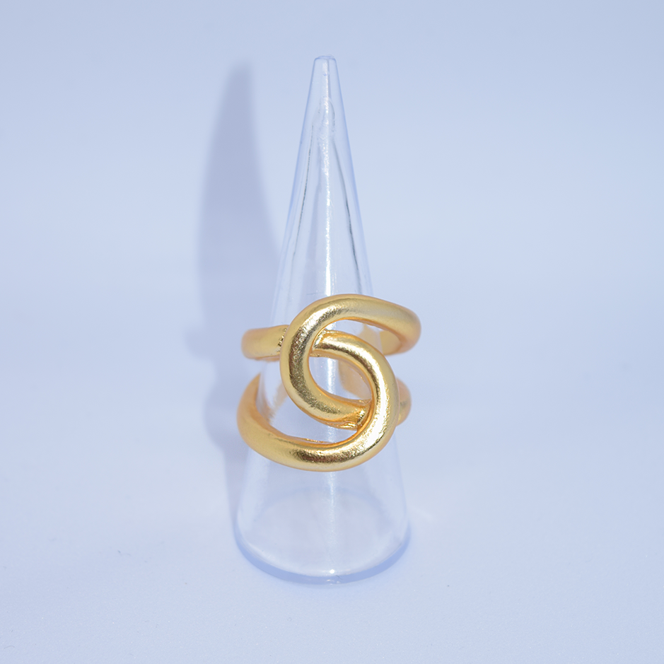 The Intertwined Link Oversize Ring, crafted in 24-kt gold with an elegant loop design, is showcased on a clear, cone-shaped stand. Its polished finish subtly reflects the light. The soft blue background enhances the ring's metallic sheen as a faint shadow is cast by the stand.