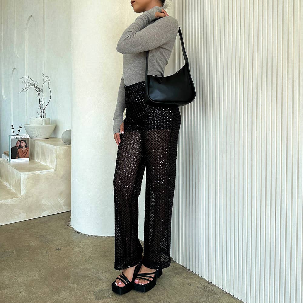 A woman in a gray long-sleeve top and black sheer pants stands by a textured white wall, holding a stylish Willow Recycled Vegan Shoulder Bag. She wears platform sandals, surrounded by white ceramic pots, a small branch, and a framed photo on the stepped concrete floor display.