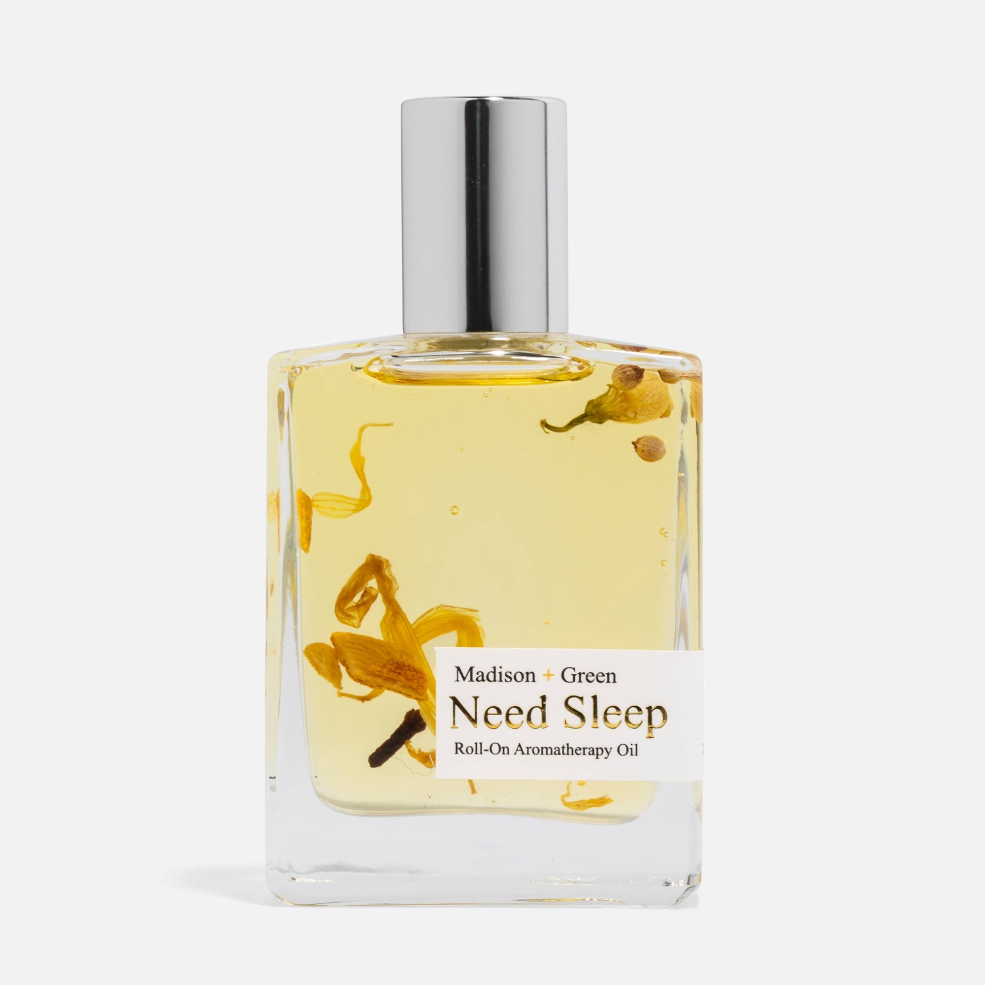 A rectangular glass bottle holds a therapeutic, stress-relieving body oil with yellow color and floating flower petals. It features a shiny silver cap and a label that reads "Need Sleep - Aromatherapy Body Oil by Madison + Green," all set against a simple white background.