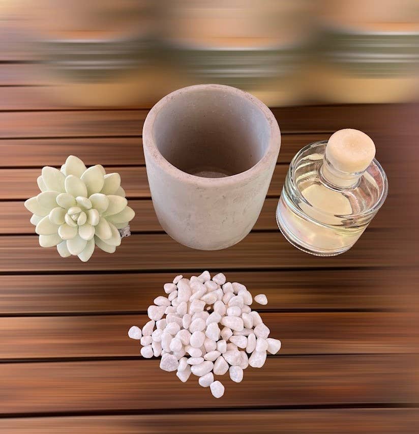 A top-view image displays the "Succulent in Planter Diffuser Gift Set - Sweet Orange Cedar," featuring a small succulent diffuser, an empty gray planter, a collection of white pebbles, and a small glass bottle with a cork stopper, all artfully arranged on a wooden surface. The light green succulent offers vibrant home greenery against a backdrop of blurred horizontal lines.