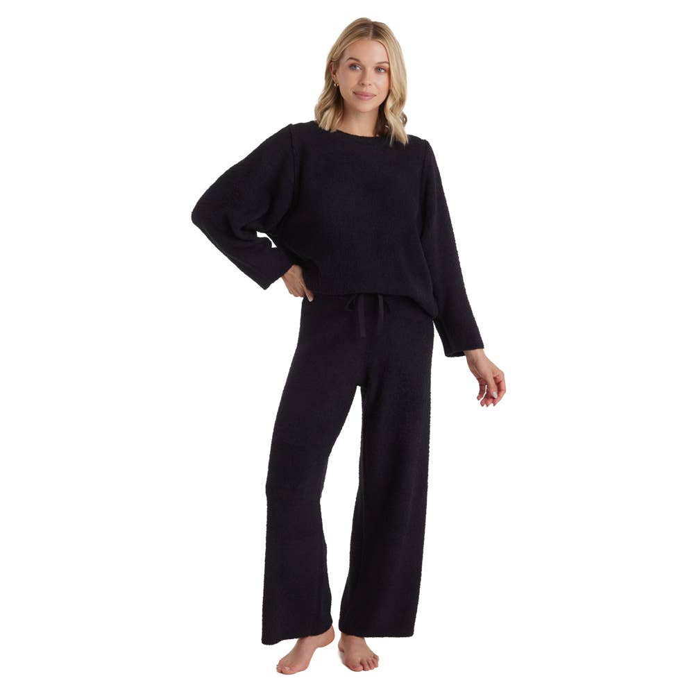 A person with long blonde hair stands barefoot, wearing the Marshmallow Lounge Set in cozy black, featuring a long-sleeve top and wide-leg pants. They pose against a plain white background, smiling softly with one hand on their hip—a true testament to Oprah’s favorite women’s lounge set.