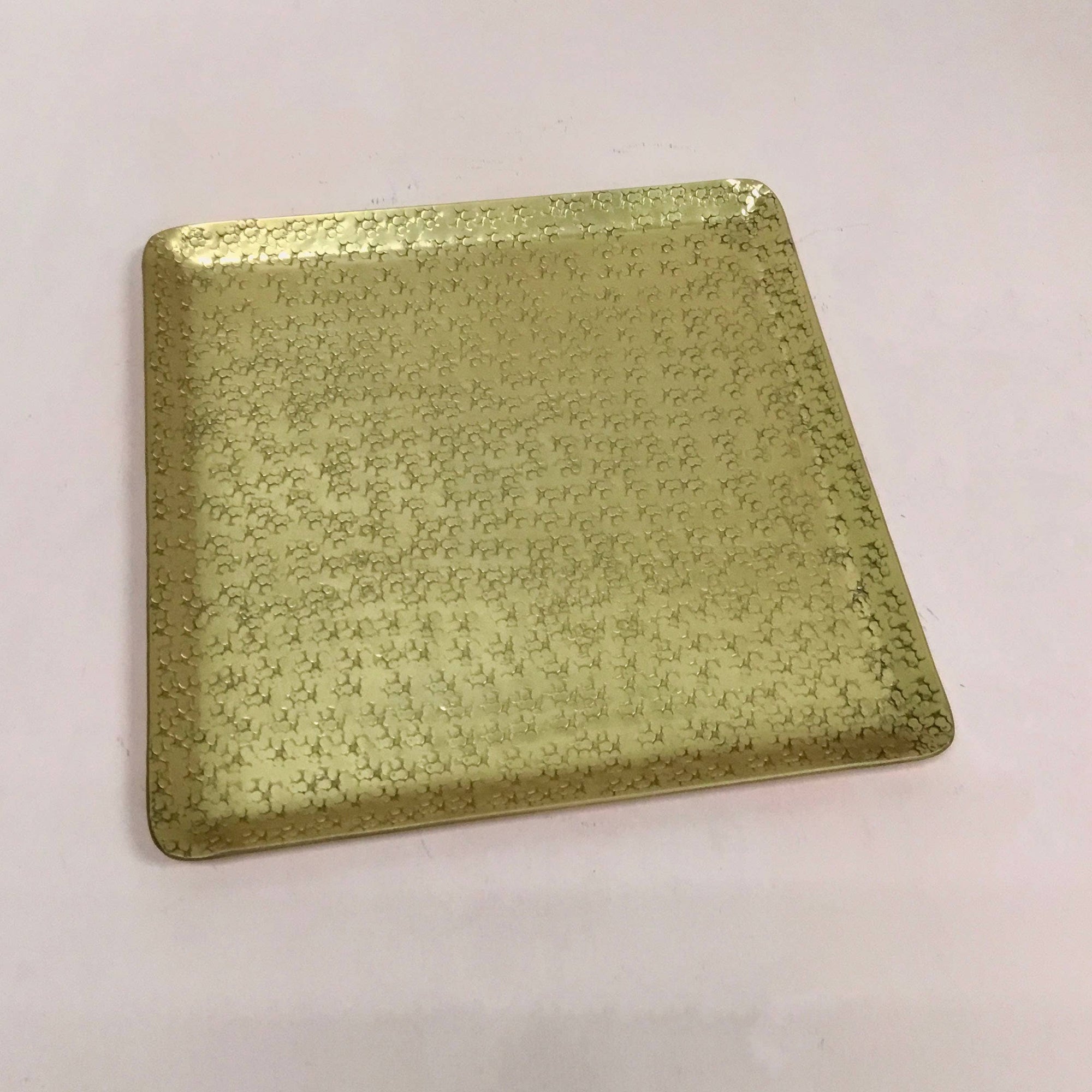 A Gold Aluminum Hammered Square Tray - 11", made from sustainable materials, rests on a smooth, light-colored surface. The elegant and refined design showcases a subtle, intricate pattern of tiny interlocking designs for added texture. Its slightly raised edges enhance its sophisticated appearance.