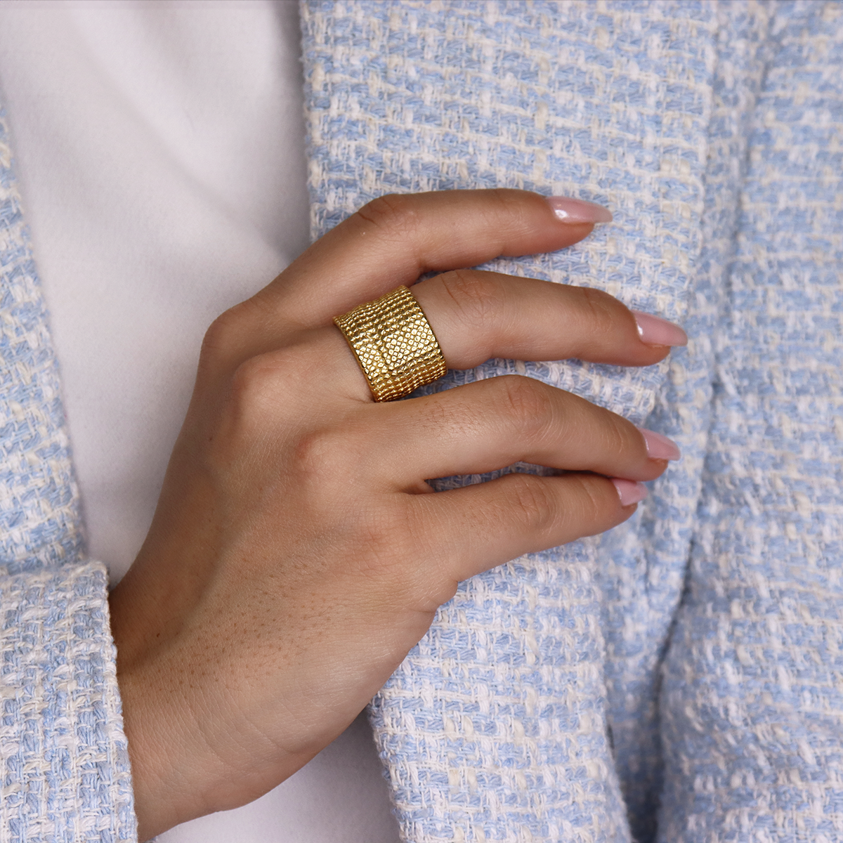 A person wearing a light blue textured jacket over a white shirt showcases their hand, which is adorned with the Chainmail Ring. The ring's 24 kt gold plating gleams brilliantly, complementing the natural, light pink polish on their fingernails. The neutral background enhances the beauty of both the hand and the ring.