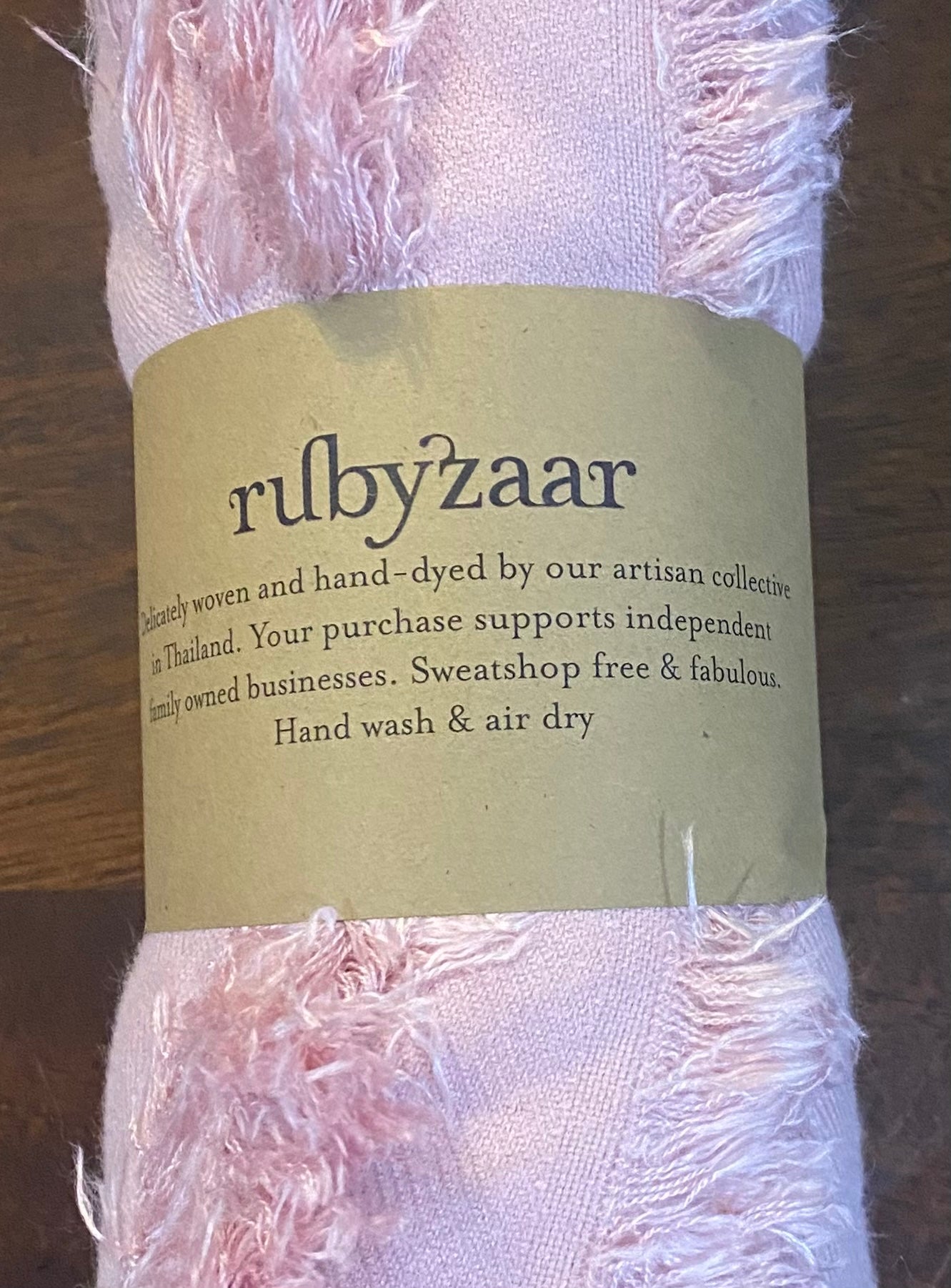 A Hima Large Scarf, Wrap, Shawl in pink features a textured fabric roll secured with a brown paper label displaying the brand name "rubyzaar" in lowercase letters. The label indicates that the fabric is hand-woven and dyed by an artisan collective in Thailand, supporting fair trade and sustainable fashion. Care instructions advise hand washing and air drying.
