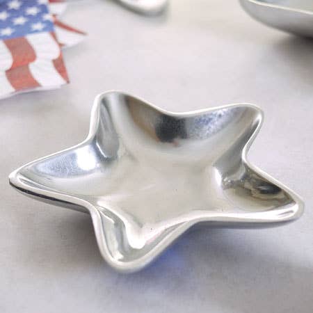 A gleaming Aluminum Star Tray rests on a light surface, with the blurred background suggesting patriotic-themed decorations featuring red, white, and blue stripes. The reflective surface of this food-safe dish captures and reflects the light, accentuating its smooth, metallic texture. For optimal results, it's recommended to hand wash.
