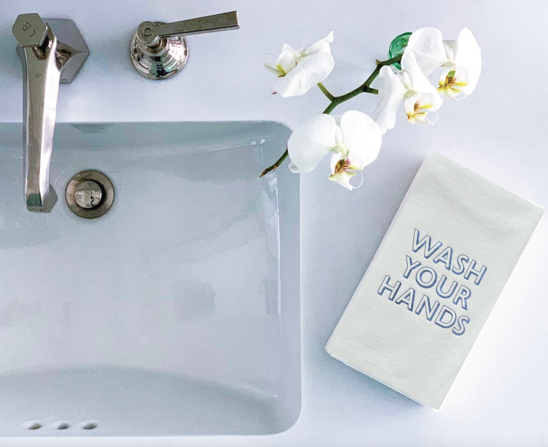 A white bathroom sink with a chrome faucet sits beside a white orchid in a vase. To the right is a clean stack of Cloth-Like Guest Towels featuring "WASH YOUR HANDS" foil-embossed on top, creating a bright, hygienic, and inviting atmosphere.