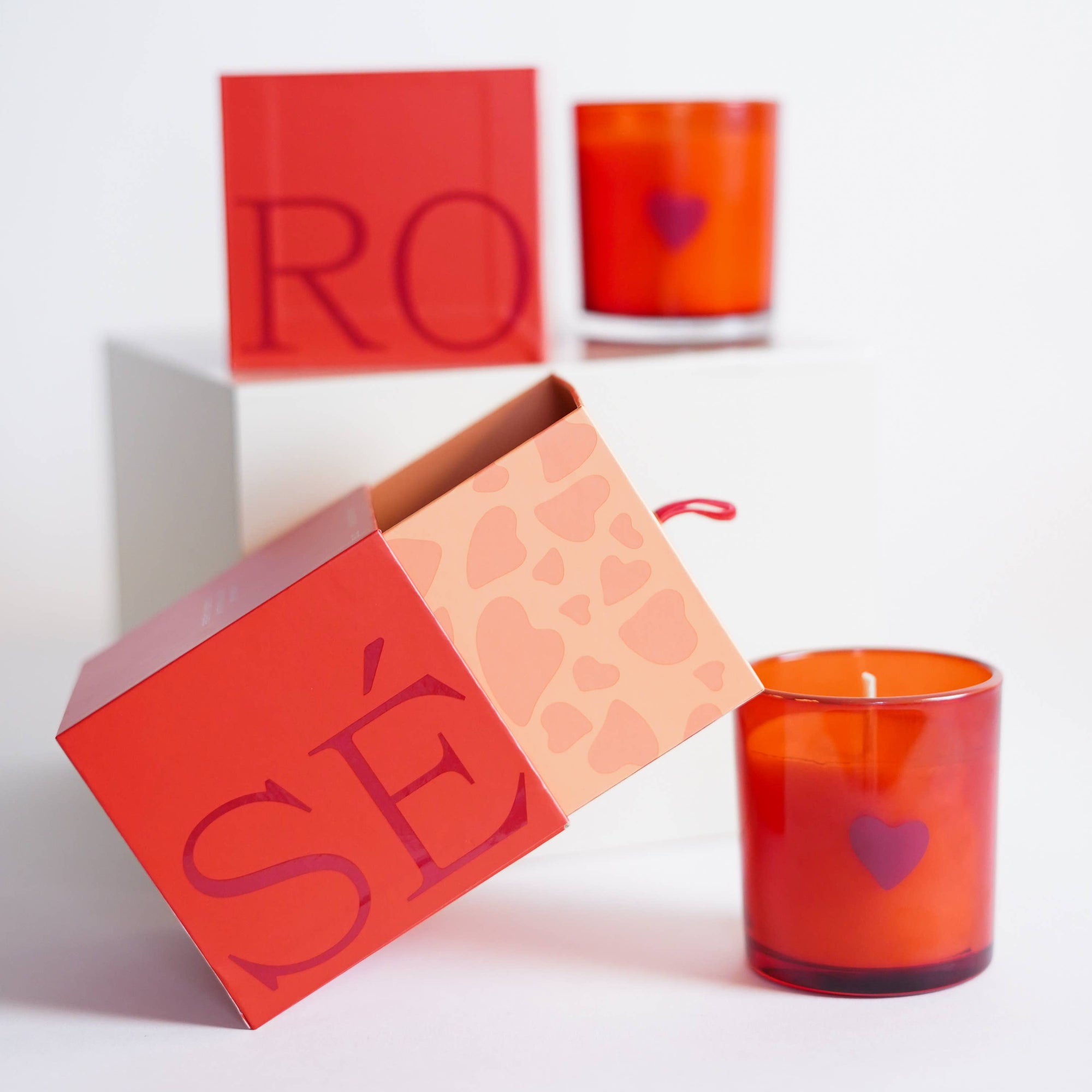 A Rosè Boxed Tumbler with a heart-adorned red glass candle sits next to an open red box with pink hearts, revealing "SÉ." In the background, a similar candle and limited-edition box showcasing "RO" suggest a delightful Rosé fragrance, all on a white surface.