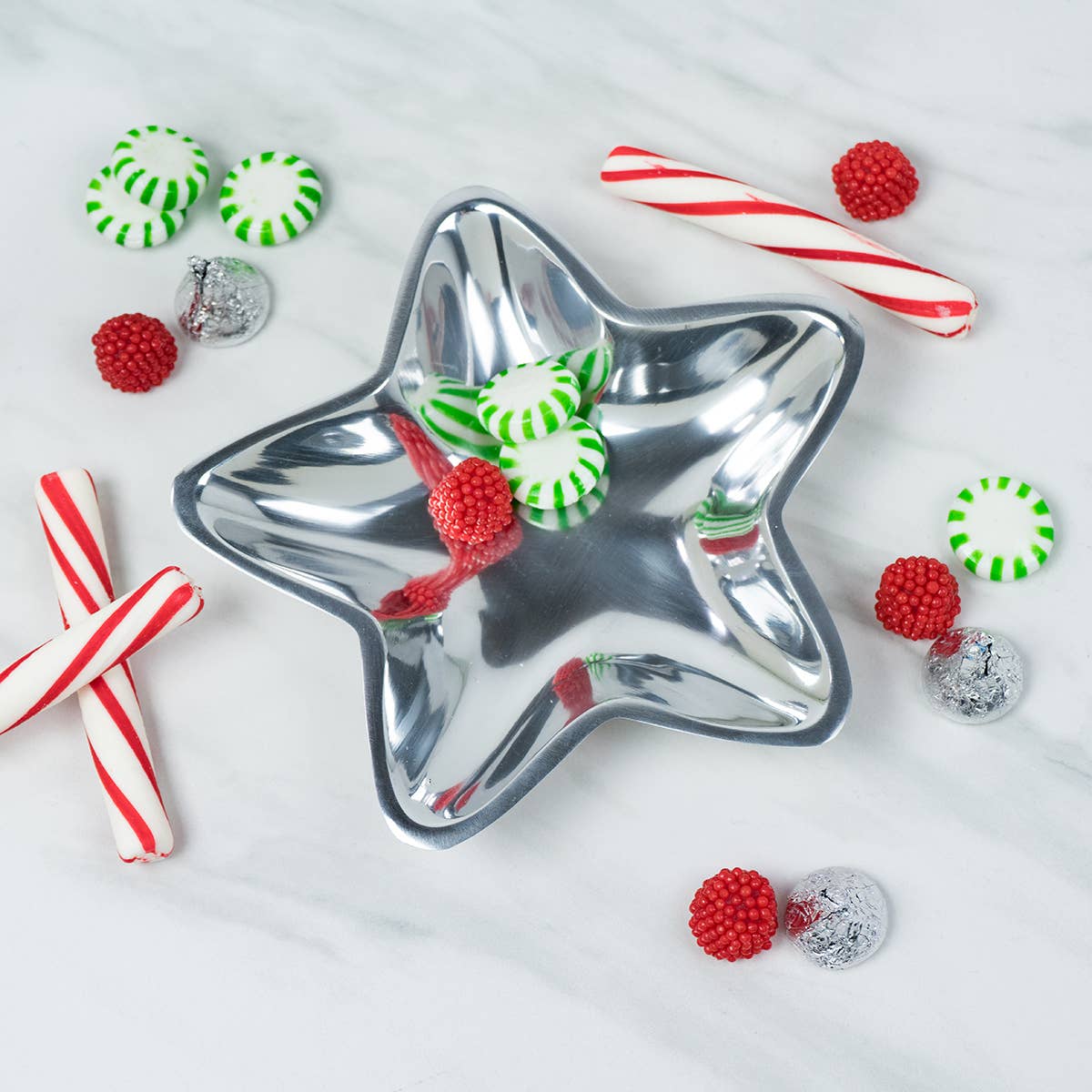 On a marble surface, the Aluminum Star Tray shines brightly, filled with red and green striped peppermints. This food-safe dish, which should be hand washed only, is encircled by festive candy canes, red gummy raspberries, and silver foil-wrapped chocolates.
