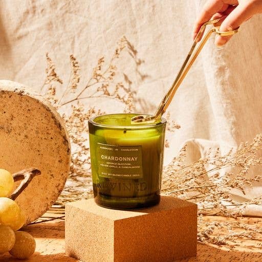 A hand uses a silver wick trimmer on the Chardonnay Candle 6 oz, made from a recycled bottle, from the Signature Collection. The green candle is on a brown block surrounded by grapes, textured stone, and dried grasses. Warm sunlight casts soft shadows to create a cozy atmosphere.