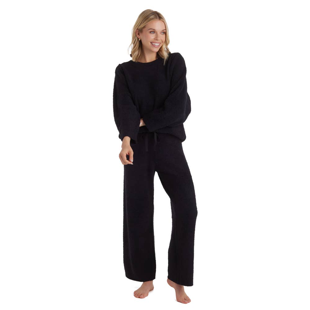 A woman stands barefoot against a white background, wearing the Marshmallow Lounge Set, an Oprah's favorite ensemble featuring a black long-sleeve fuzzy sweater and matching wide-leg pants. With shoulder-length blonde hair, she's smiling while looking to her right. Her hands rest at her sides, and her left foot is slightly forward.