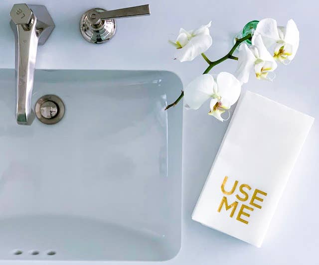 A bathroom sink with a metallic faucet on the left. To the right, a luxurious towel from the Cloth-Like Guest Towels - Year Round Themes collection showcases "USE ME" in gold lettering. Above the towel, a green vase holds white orchids, resting on a glossy white countertop.