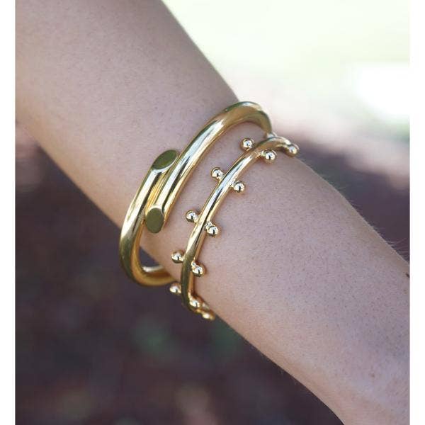 A close-up of a person's arm highlights two gold bracelets. One features a smooth design with rounded ends, while the Cindy Studded Cuff is adorned with small spherical accents along its curve. Both showcase an 18k gold-plated finish and hypoallergenic design, gleaming against softly blurred backgrounds and emphasizing their elegance.