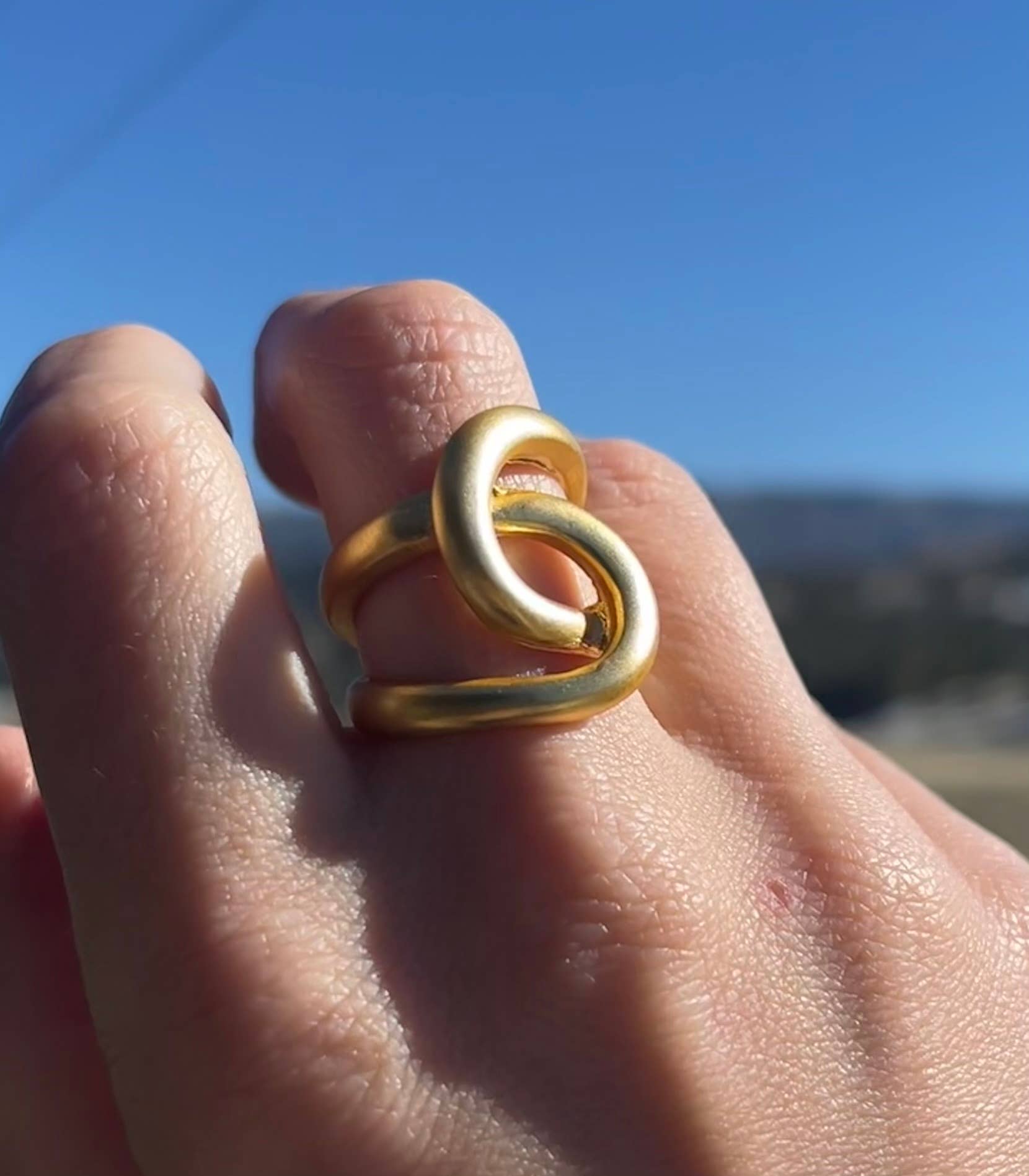 A close-up of a hand adorned with the eye-catching Intertwined Link Oversize Ring, crafted from 24-karat gold and showcasing an abstract, intertwined design, set against a clear blue sky. The ring's polished surface catches the sunlight beautifully, highlighting its elegant curves. In the blurred background, an outdoor landscape is faintly visible.