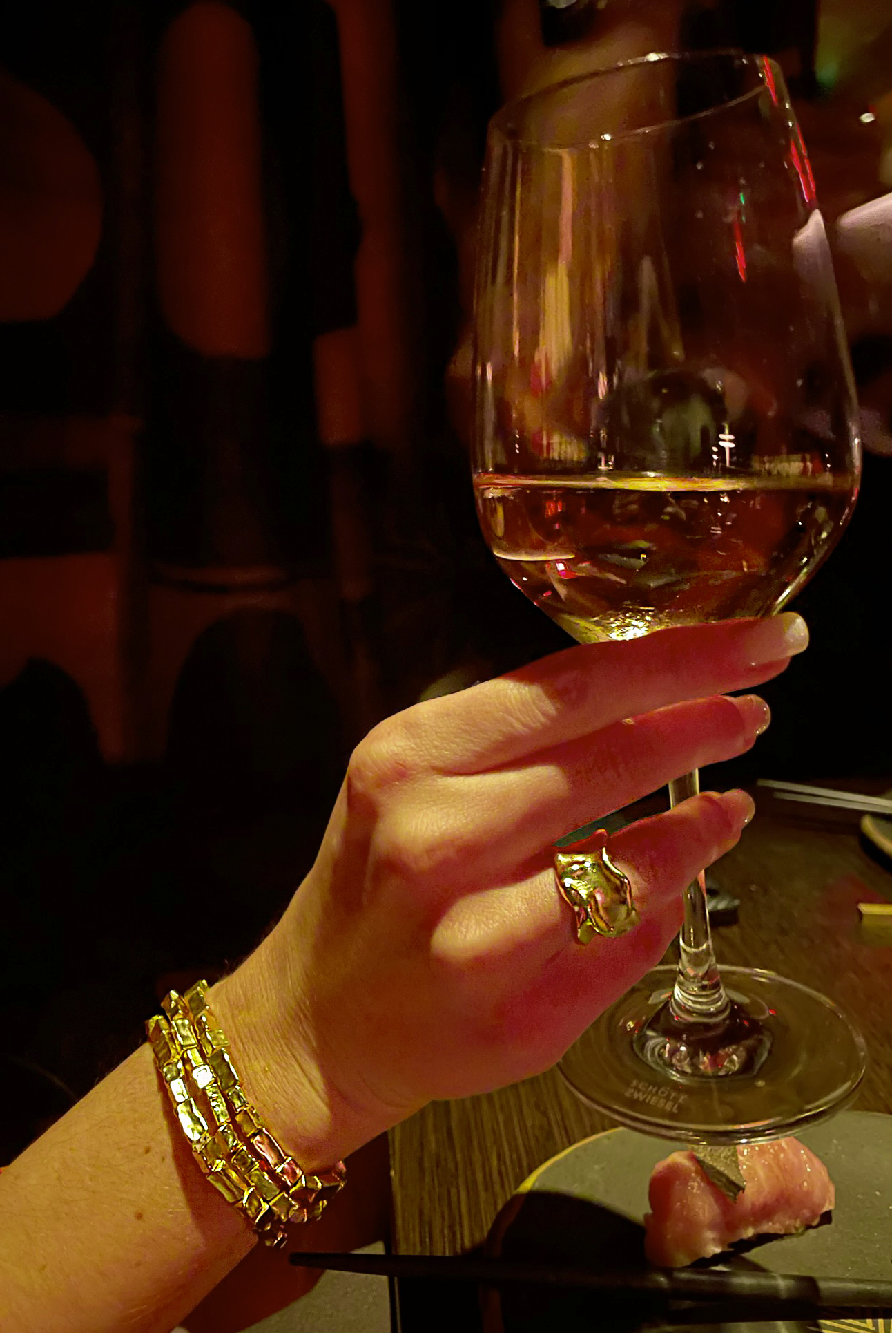 A hand adorned with the adjustable Crumpled Foil Ring and gold-plated bracelets holds a glass of white wine. The background is dimly lit, setting a cozy atmosphere. A piece of sushi rests on a nearby plate, with chopsticks elegantly placed on the dark table.