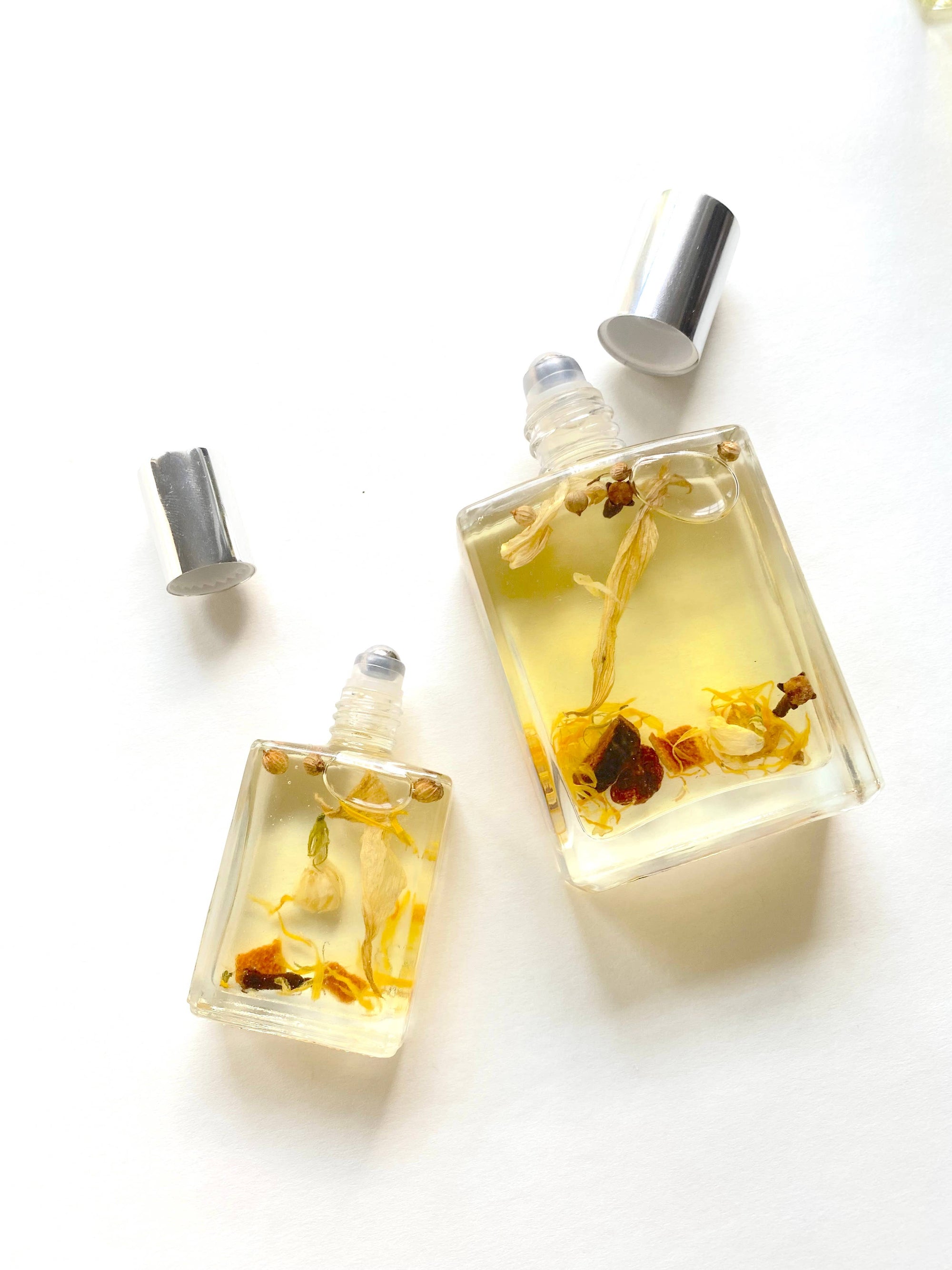 On a white surface, two glass bottles with silver caps rest, each filled with "Need Sleep" - Aromatherapy Body Oil. The larger bottle showcases floating dried flowers and herbs for stress relief, while the smaller one reflects the same elegant design with similar floral inclusions. The caps are placed next to the bottles.