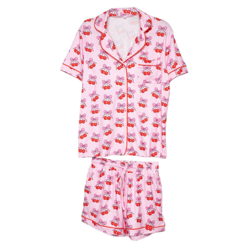 This Cherry Bow Cotton Pajama set includes a pink, short-sleeve, collared button-down top with red piping and matching elastic waistband shorts. Red cherries with green stems in a playful bow print enhance the summery feel.