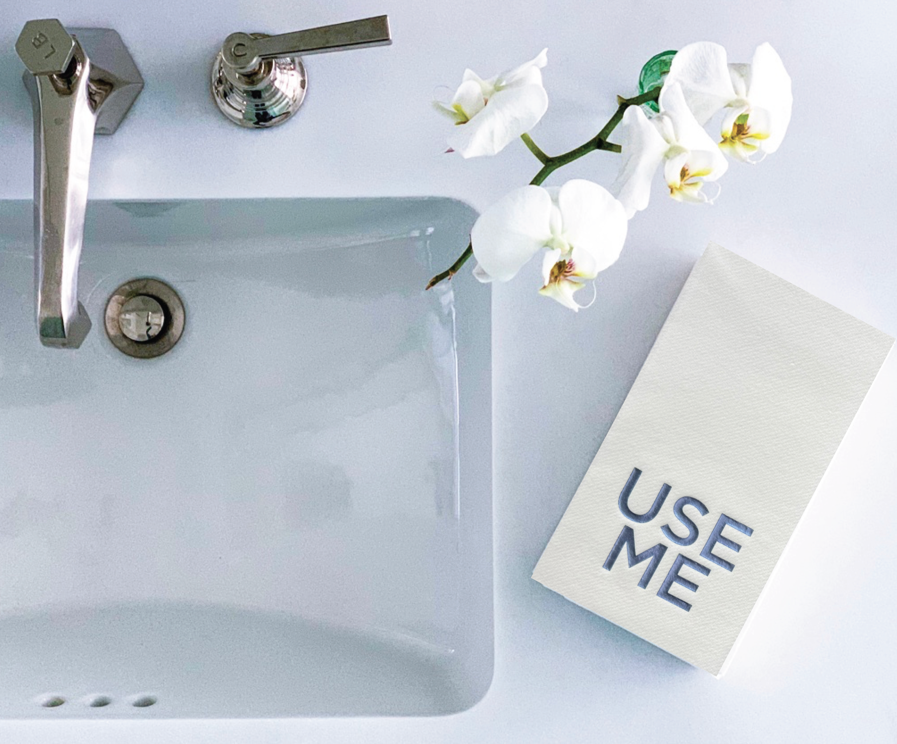 A modern bathroom sink with a silver faucet is accompanied by white orchid flowers. To the right, a stack of Cloth-Like Guest Towels - Year Round Themes graces the counter, featuring "USE ME" printed in bold black letters. The arrangement presents a clean and minimalist aesthetic.