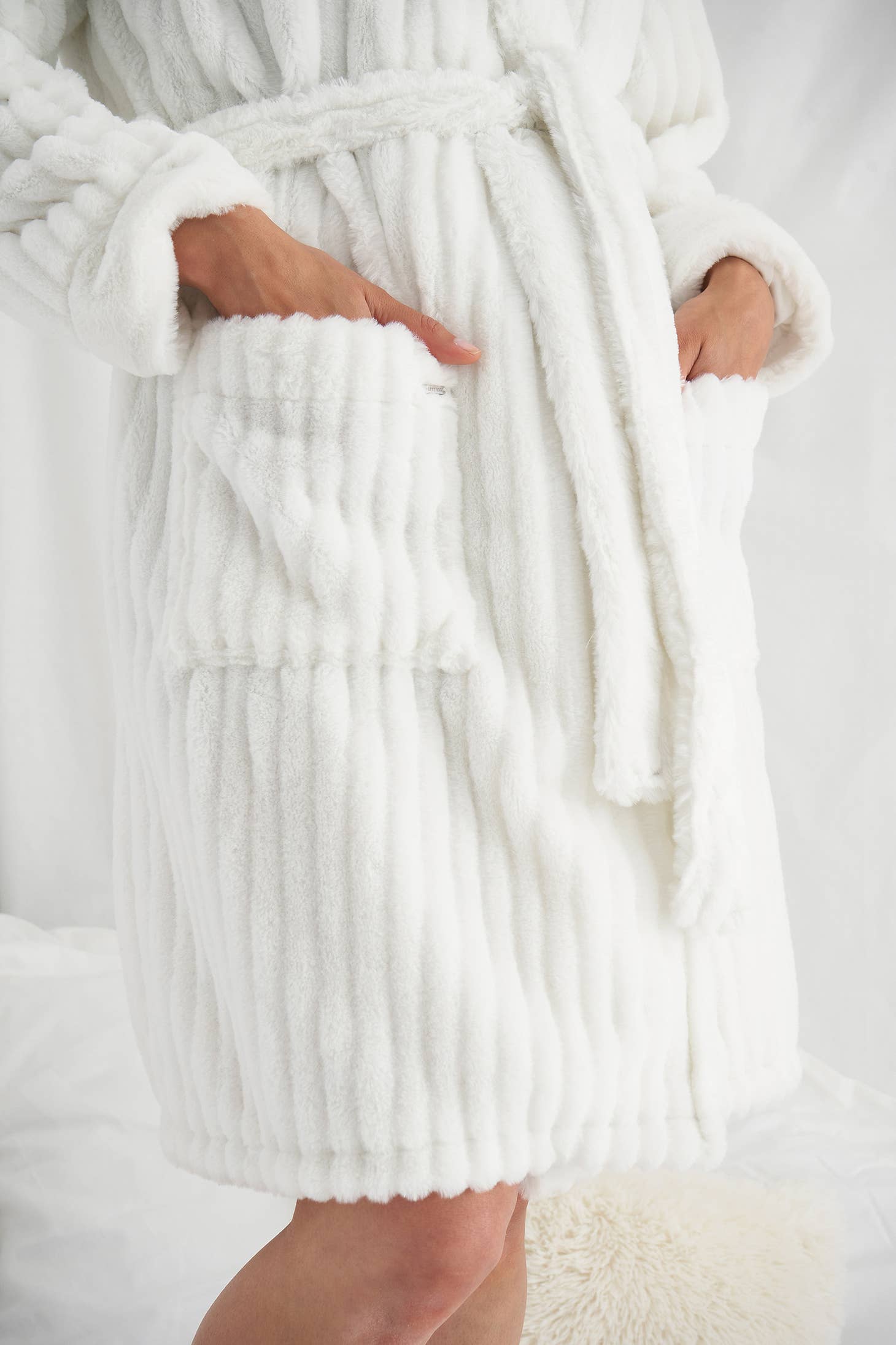 Dressed in a white Cloud Robe characterized by its textured plushness, an individual is photographed from the shoulders down. Their hands are gently tucked into the robe's deep front pockets, while a coordinating belt cinches at the waist. The softly blurred background enhances a cozy and tranquil ambiance.