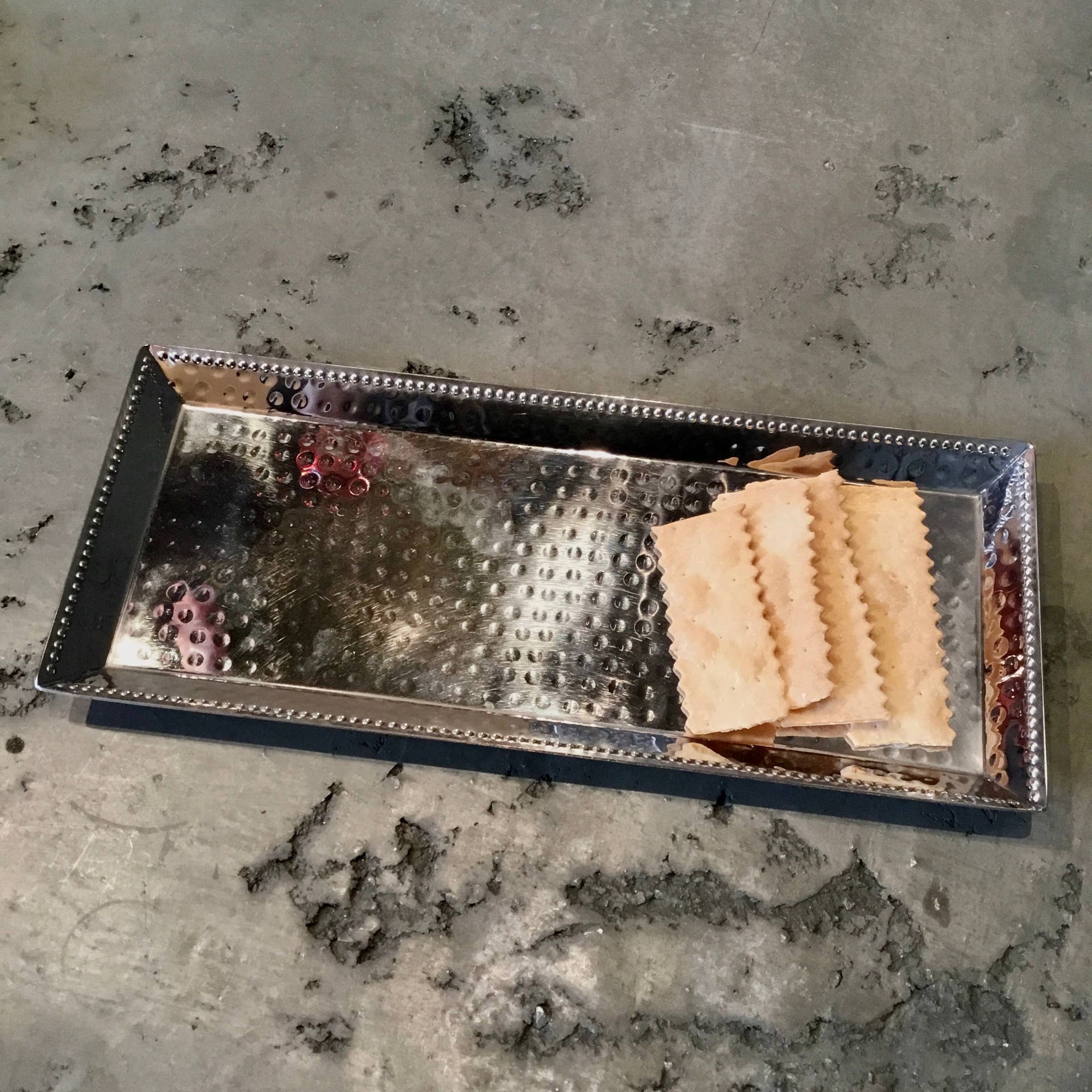 A Hammered Stainless Steel Rectangle Tray w/ Beaded Edges sits on a gray, marbled surface, featuring a textured design and five neatly stacked rectangular crackers on the right side. The tray's slightly raised beaded edges and polished finish subtly reflect the light.