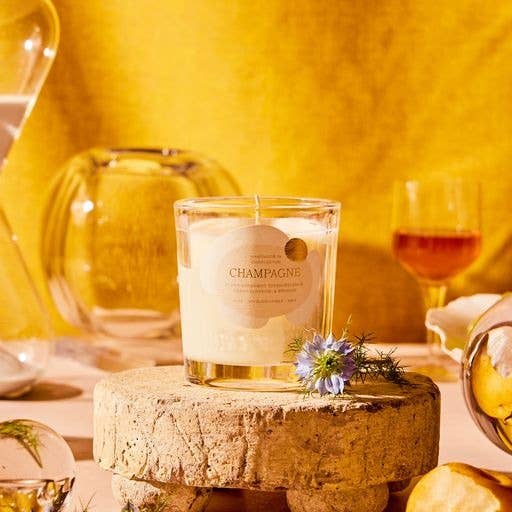 A 6 oz Champagne Candle from the Sparkling Collection sits on a rustic wooden stand adorned with small flowers. A glass decanter and wine glass with amber liquid add elegance against a warm yellow wall. Soft lighting makes it ideal for any celebratory occasion.