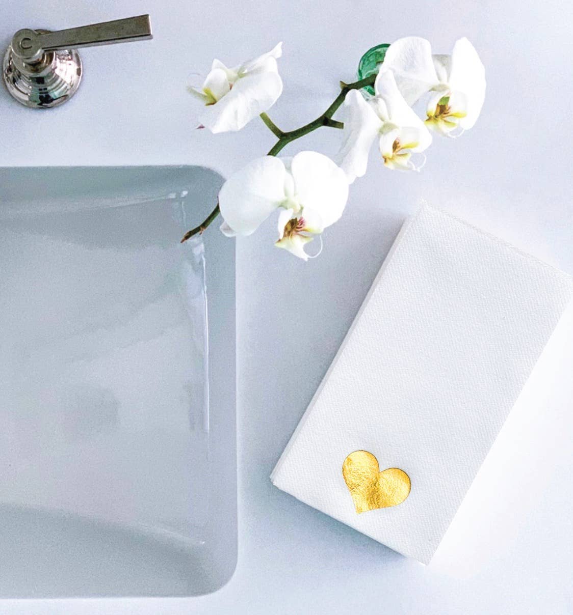 A white orchid branch rests on a light gray countertop next to a sink with a metallic faucet. Nearby, Cloth-Like Guest Towels featuring a gold heart design add elegance, making them perfect as bathroom napkins.
