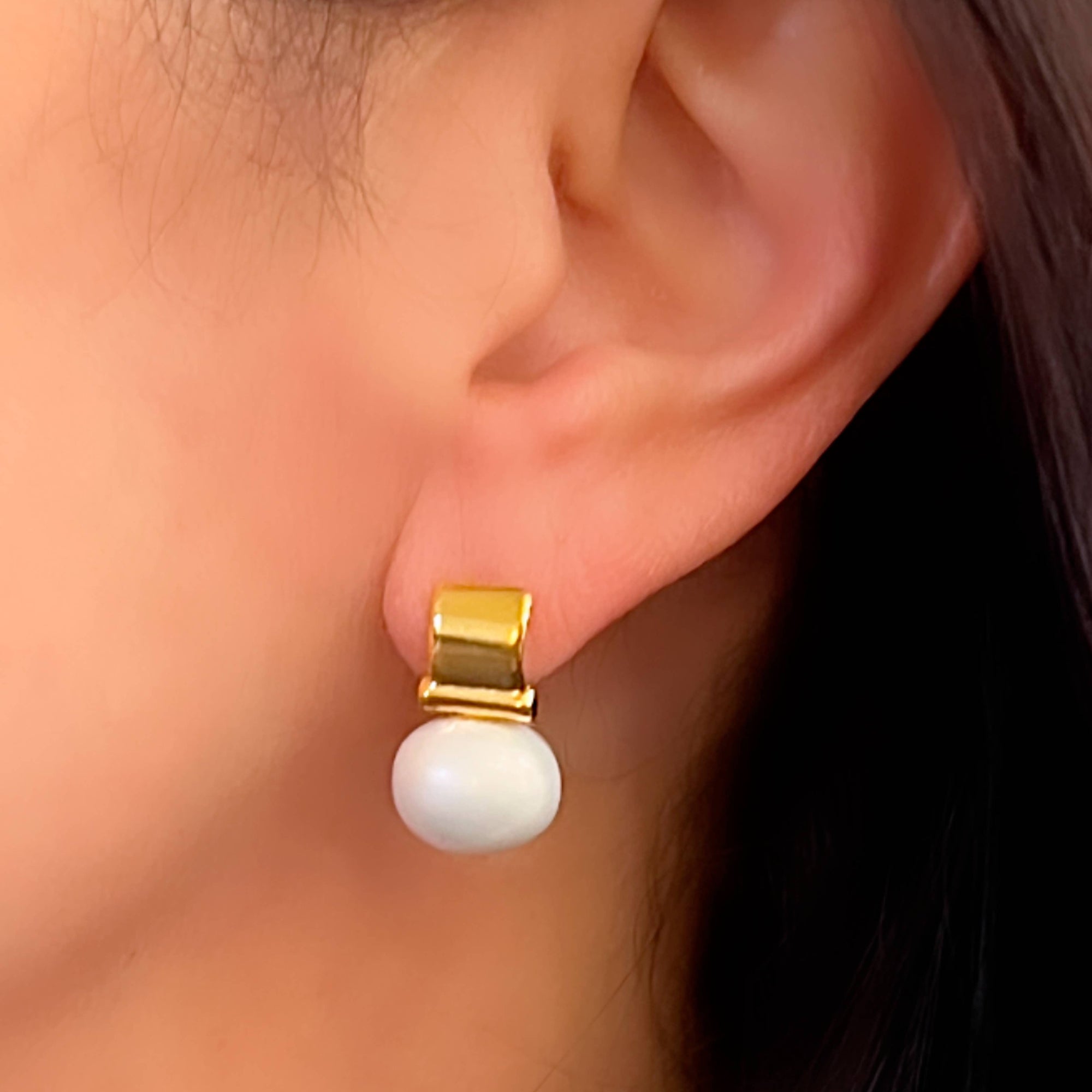 A close-up captures an ear wearing the Smooth + Classic Pebble Pearl Post Earrings, made from 14 karat gold and showcasing a white pearl. The rectangular gold section elegantly complements the pearl's dangle. Soft lighting accentuates its sheen, while hypoallergenic backing ensures comfort against hair and skin in the background.