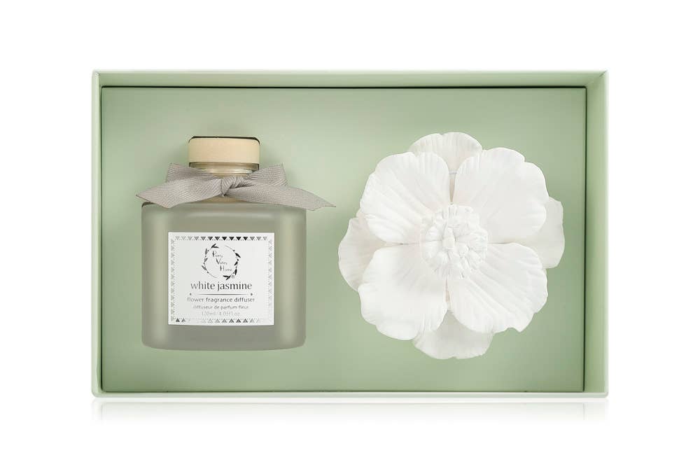 A sophisticated green gift box beautifully holds a frosted glass bottle labeled "White Jasmine," accentuated with a beige bow. Accompanying it is an intricately designed large white ceramic flower decoration. The Marigold Ceramic Flower Diffuser Gift Set - White Jasmine rests against a soft green background, radiating charm and elegance.