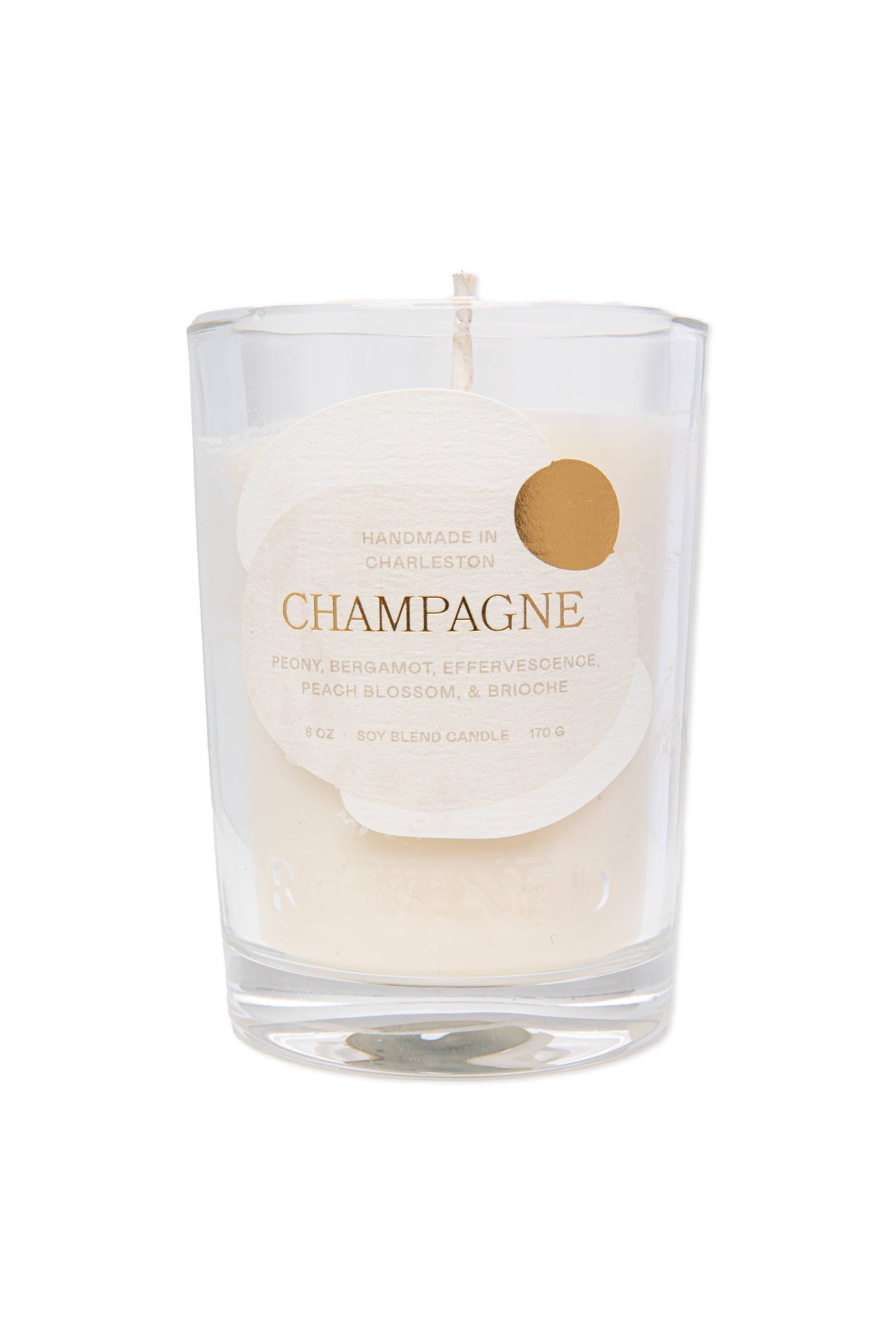 The Champagne Candle 6 oz from the Sparkling Collection is a clear glass candle with a circular tag listing scents: peony, bergamot, effervescence, peach blossom, and brioche. Handmade in Charleston, this minimalist white candle enhances any celebratory occasion.