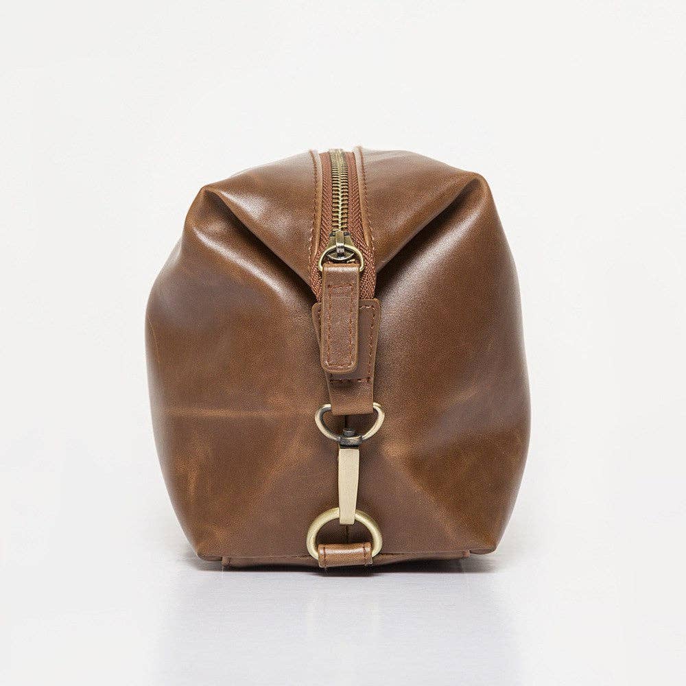 The Journeyman Dopp Kit, crafted from brown vegan leather and accented with a gold zipper and metal clasp, is showcased upright. Its smooth and polished surface exudes a rich, warm tone. The design includes a central seam, an easy-to-handle loop, and offers ample space for grooming essentials.