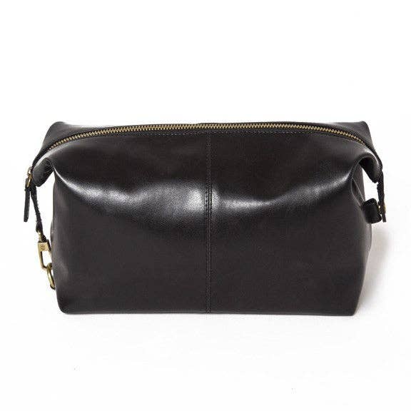 The Journeyman Dopp Kit is a stylish black vegan leather bag featuring a gold zipper closure running along the top. It has a structured rectangular shape with slightly folded corners and subtle stitching on the front. For easy handling of your grooming essentials, small loops are attached to the ends of the zipper.