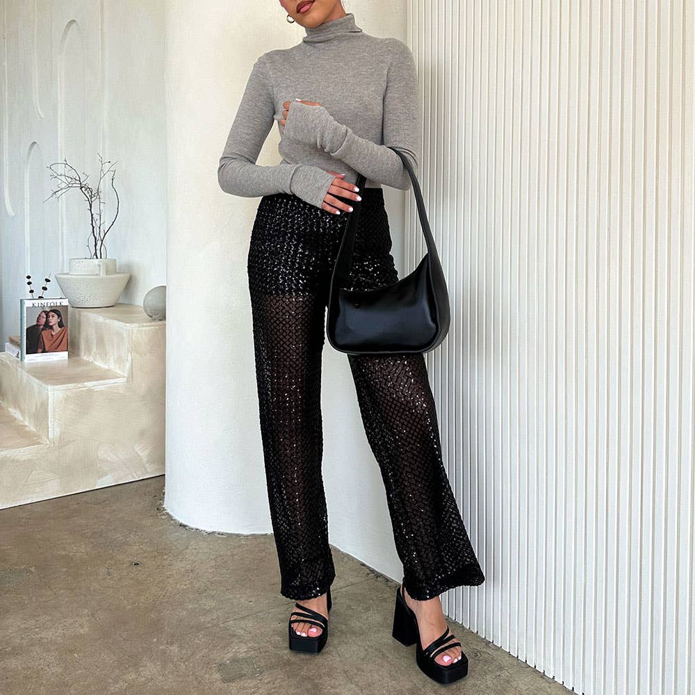 In a stylish, minimal interior with a curved wall and modern decor, someone wears a gray turtleneck, black sequined pants, platform sandals, and holds the Willow Recycled Vegan Shoulder Bag.