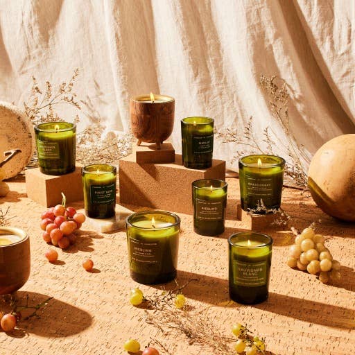 A rustic setting with Merlot Candle 6 oz in glass jars on wood, encircled by grape-like clusters of red and green, dried flowers, and textured stones. Off-white draped fabric in the background enhances a cozy ambiance ideal for wine enthusiasts.