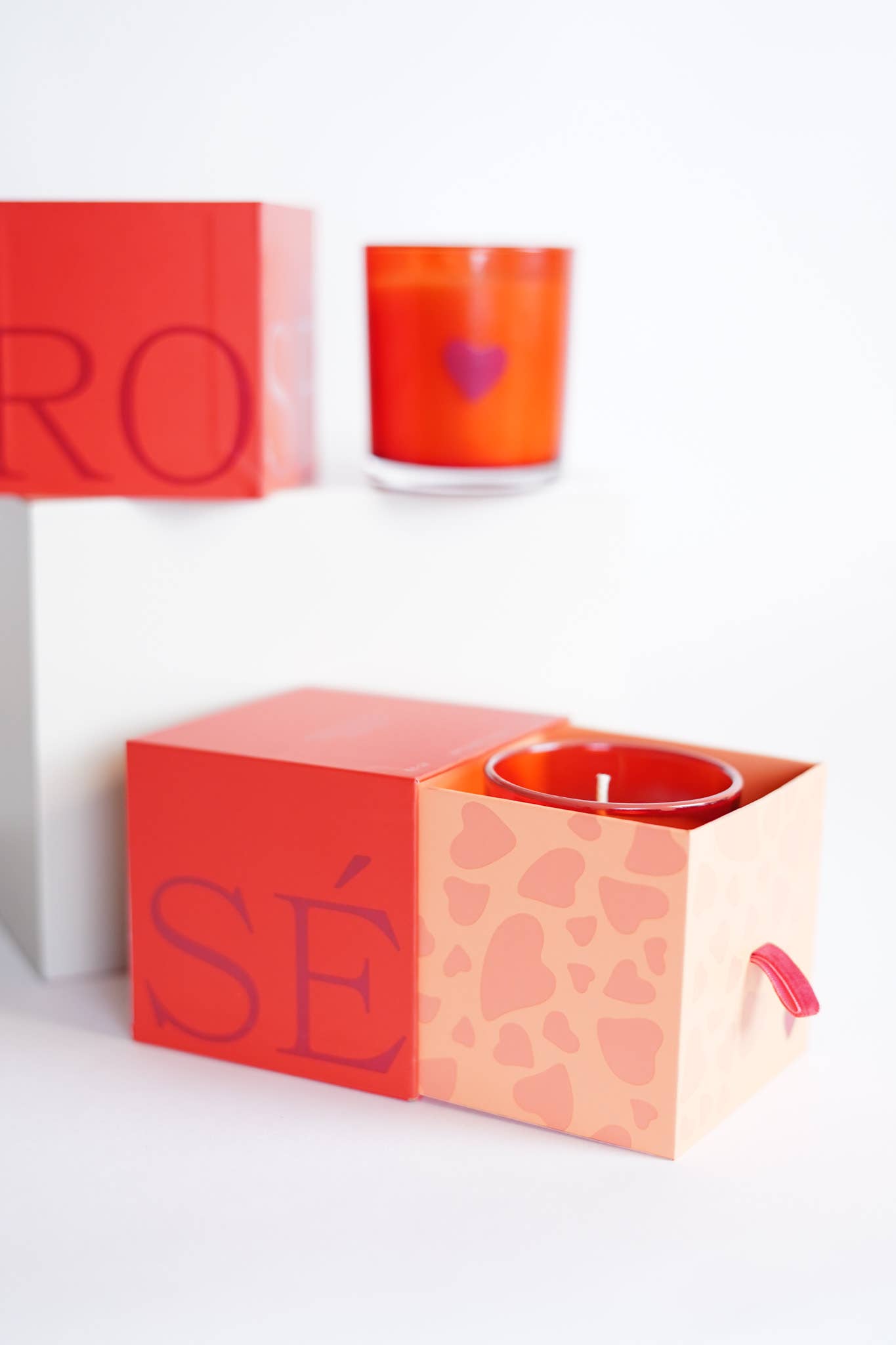 A red-orange candle with a heart design in a glass jar sits next to an open square box displaying the Rosè fragrance candle. The box, featuring pink and hearts with red sides, partially reveals the product. Another similar unopened limited-edition "RO SÉ" box is stacked on top in a white background.